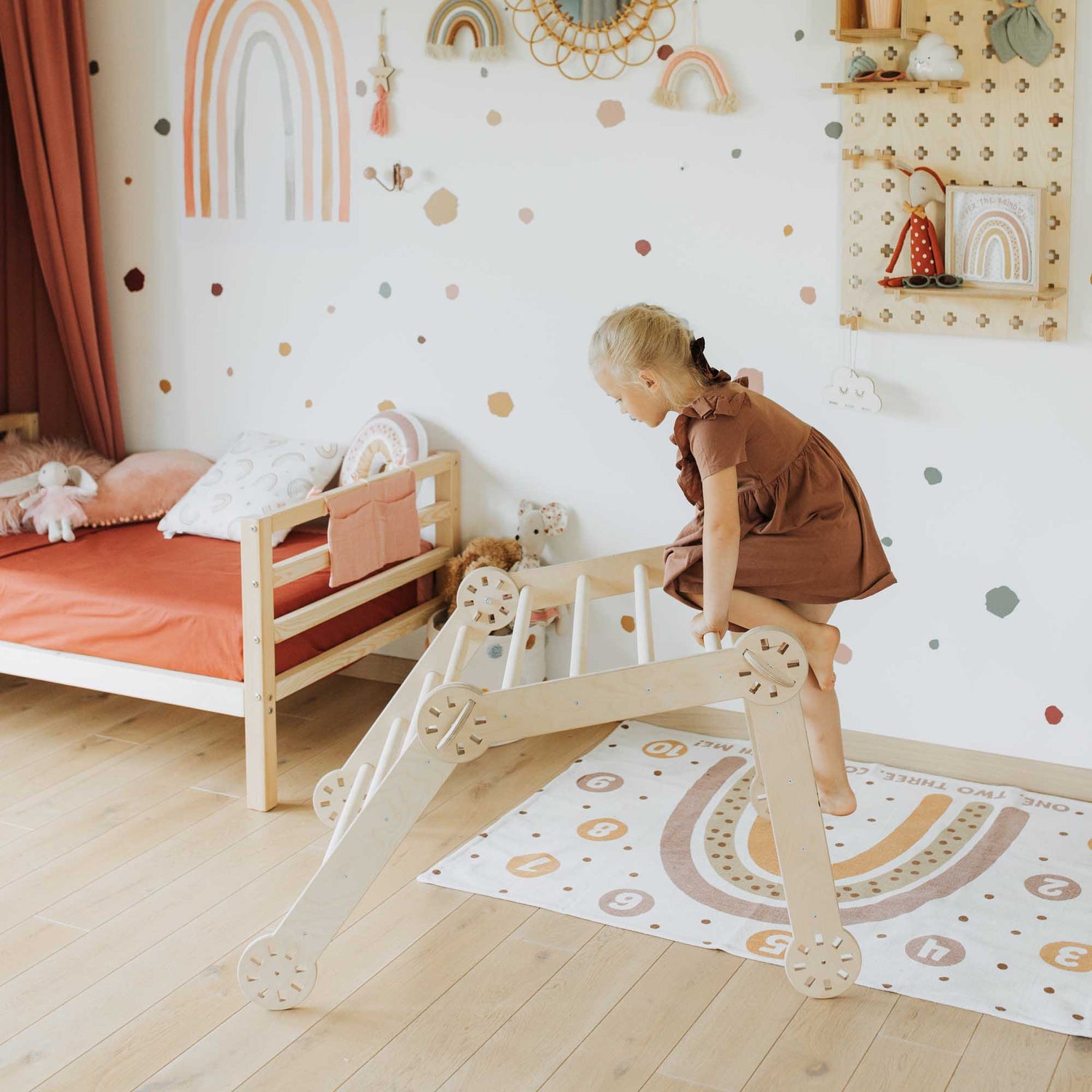 A little girl standing on a wooden ladder in a child's room, balancing on the Climbing arch + Transformable climbing gym + ramp from Sweet Home From Wood.