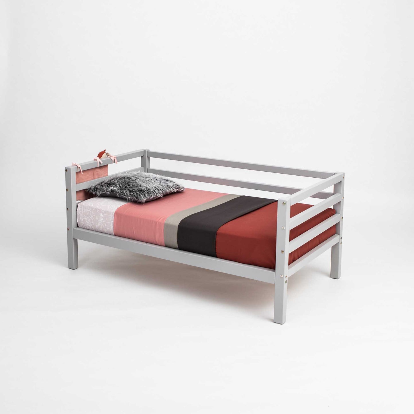 A Kids' bed on legs with a three-sided horizontal rail features a gray solid wood frame, multicolored striped bedspread, two pillows, and a plush toy, creating a cozy Montessori-inspired toddler setup against a plain white background.
