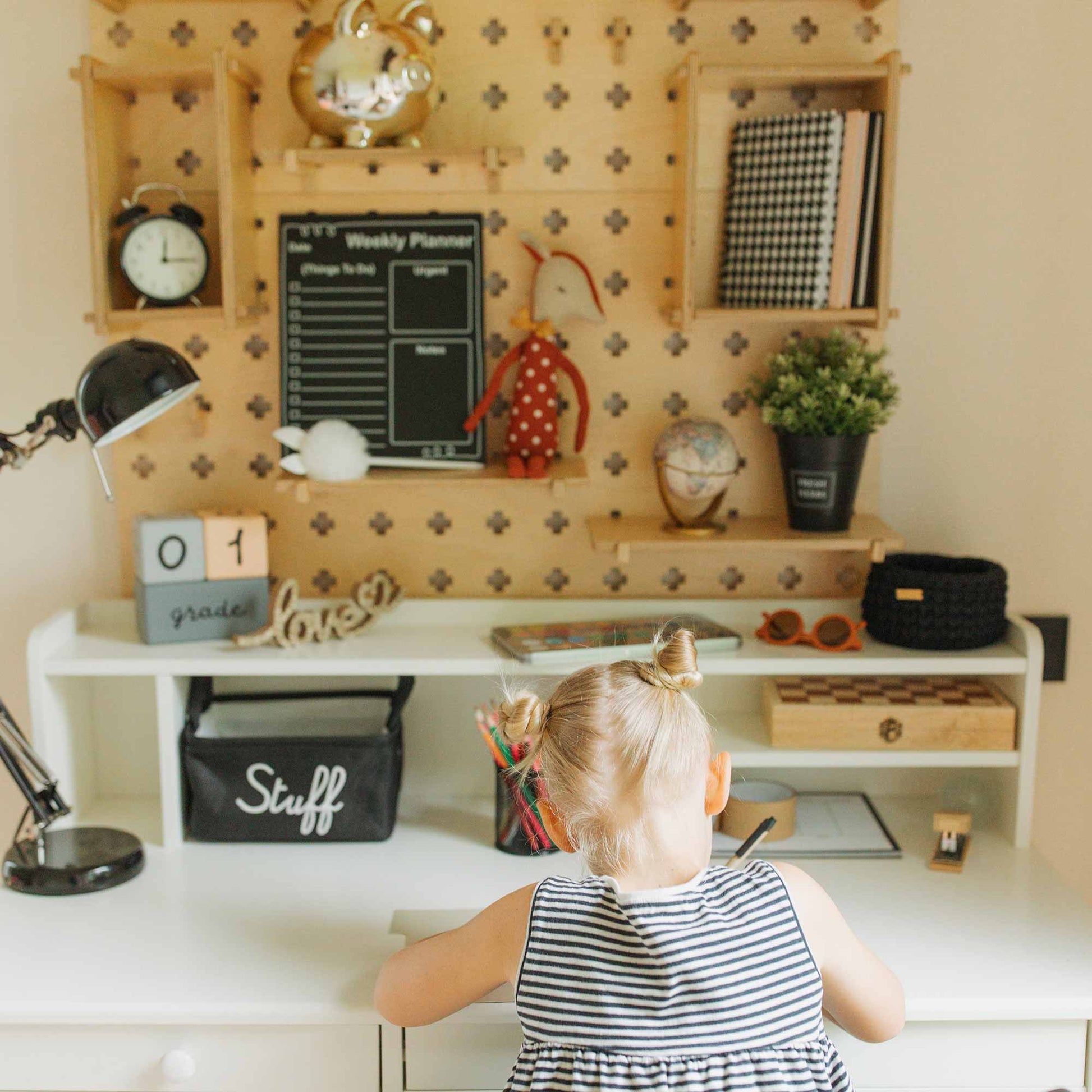 A young child in a striped dress sits at a desk, facing a decoratively arranged wall featuring the Large Pegboard Shelf with Floating Shelves, alongside a weekly planner and various items including a lamp, plant, clock, and toys.