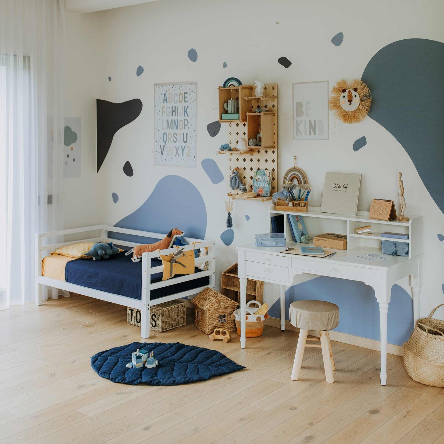 Children’s bedroom with a Kids' bed on legs featuring a horizontal rail on 3 sides, blue and white decor, desk with stool, wall shelves, baskets, wooden toys, and educational posters.