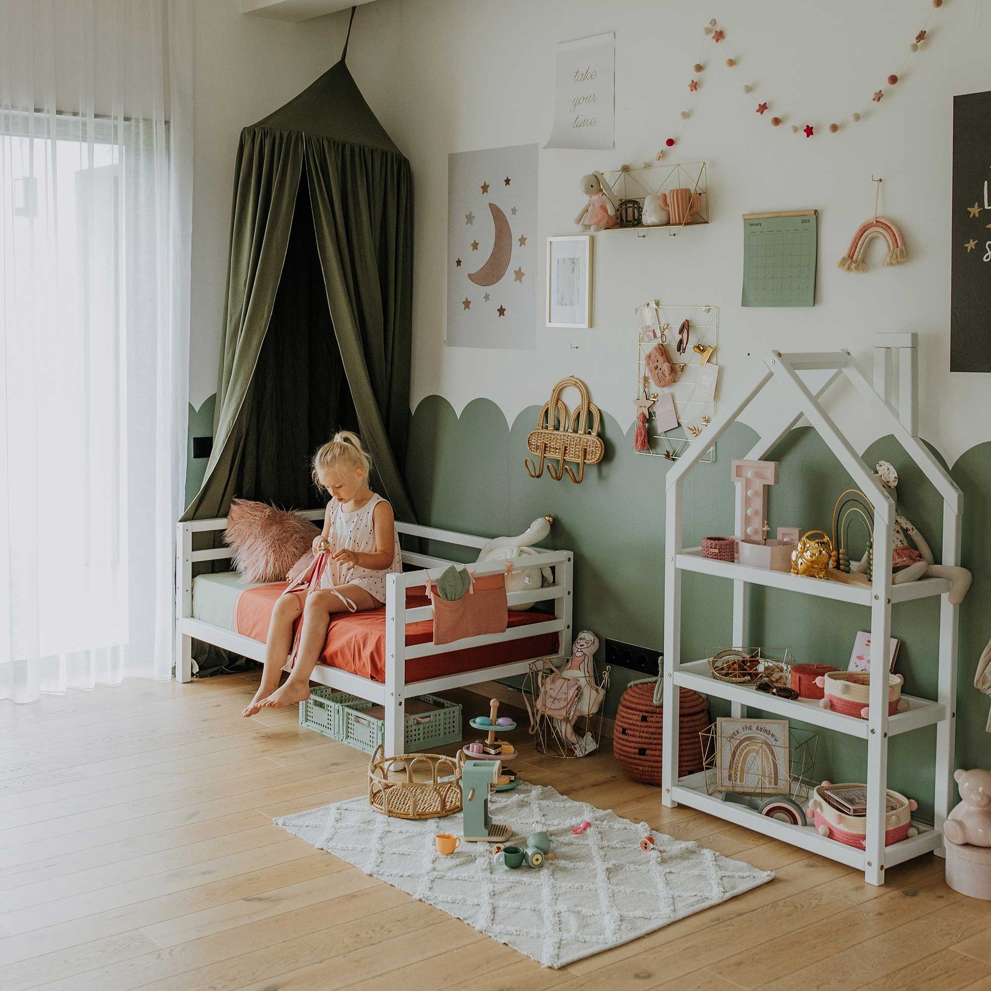 A young girl sits on a Kids' bed on legs with horizontal rail on 3-sides in a tidy children's room decorated with toys, wall art, a house-shaped shelf, and wooden floors.