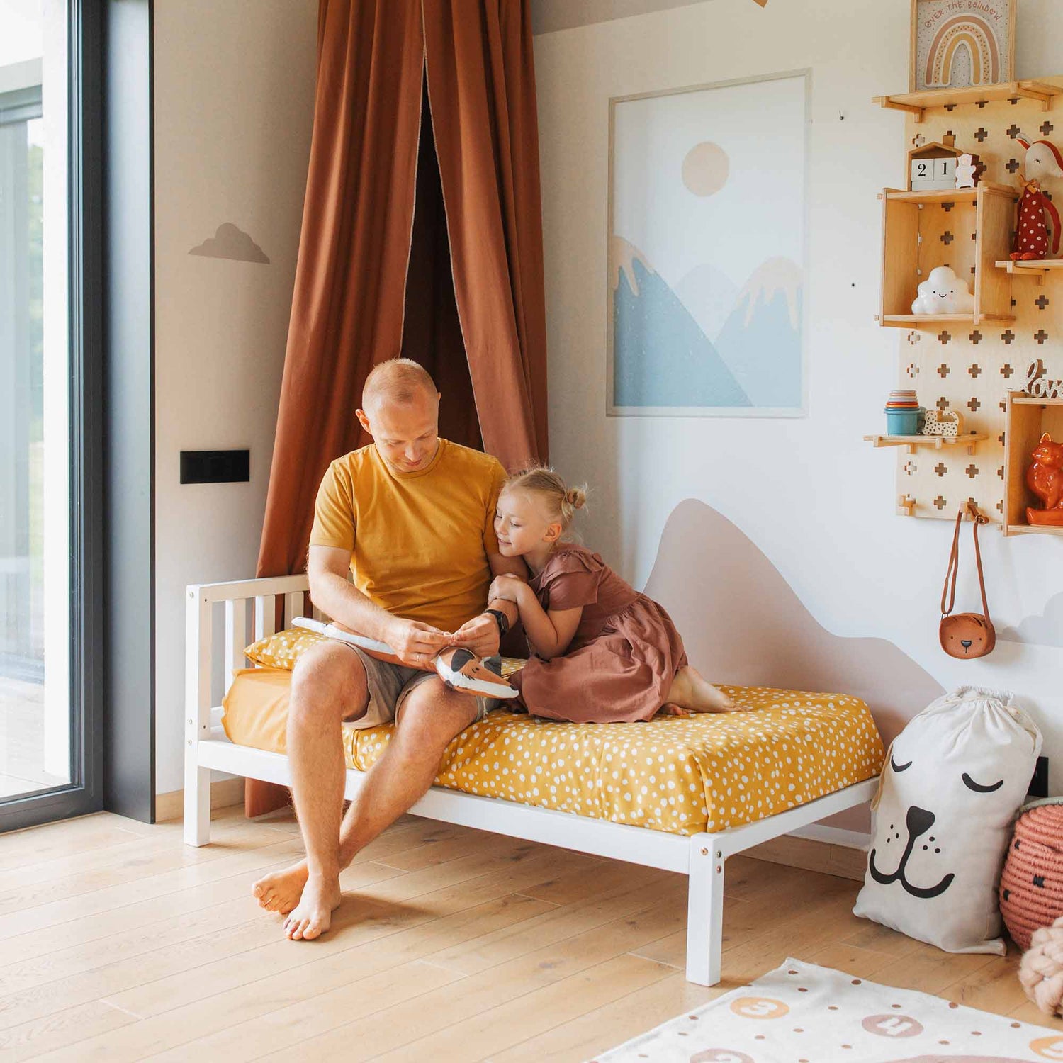 An adult helps a child put on shoes while sitting on a Kids' bed on legs with a headboard in a neatly decorated, Montessori-inspired bedroom.