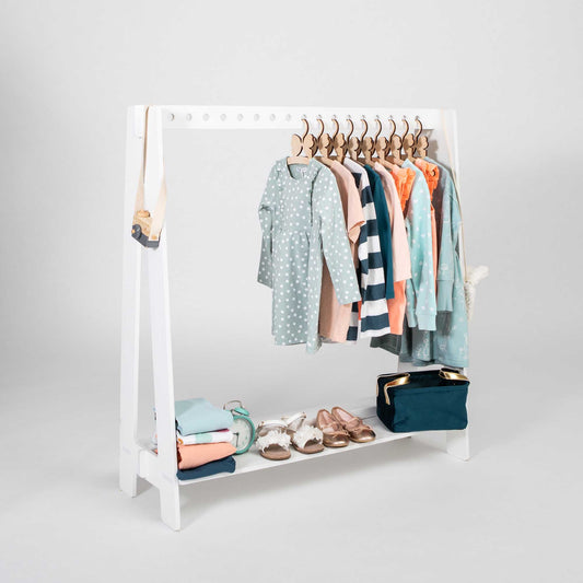 The Montessori clothing rack, a white timber structure, beautifully displays children's clothing such as dresses, shirts, and a sweater. Its bottom shelf is utilized for neatly folded clothes, shoes, a backpack, and a purse. Set against a plain gray background, this setup is ideal for efficient closet organization.