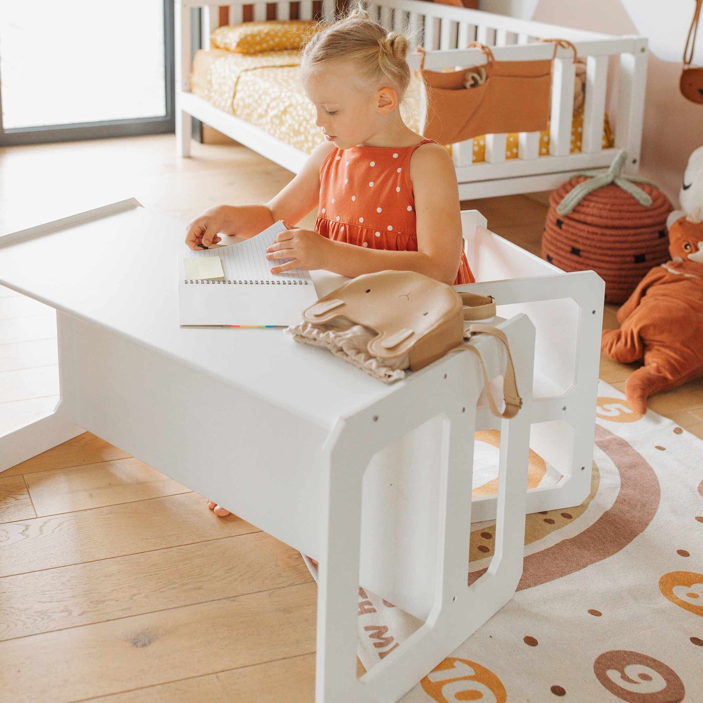A toddler in an orange dress sits at a white Montessori weaning table and chair set, drawing in a notebook. A small animal-shaped bag is on the desk. A crib and plush toys are in the background on a wooden floor, creating a Montessori-inspired setting that fosters fine motor skill development and encourages toddler independence.