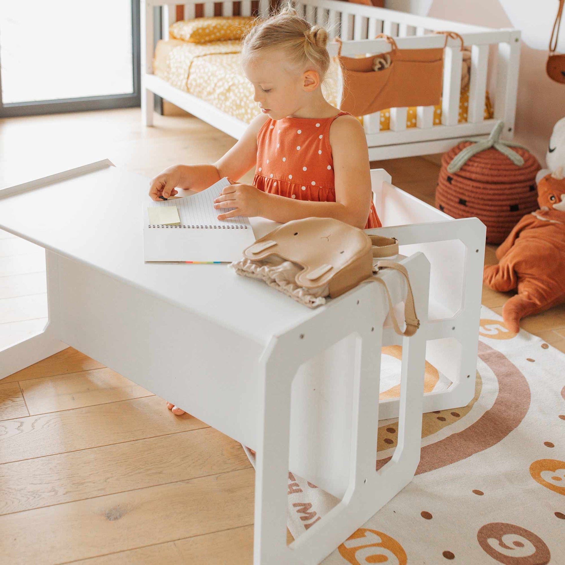 A toddler in an orange dress sits at a white Montessori weaning table and chair set, drawing in a notebook. A small animal-shaped bag is on the desk. A crib and plush toys are in the background on a wooden floor, creating a Montessori-inspired setting that fosters fine motor skill development and encourages toddler independence.