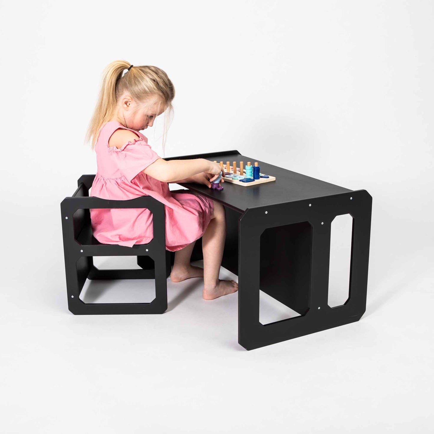 A young girl with a ponytail in a pink dress sits at a Montessori weaning table and chair set, playing with a small chessboard, fostering fine motor skill development.