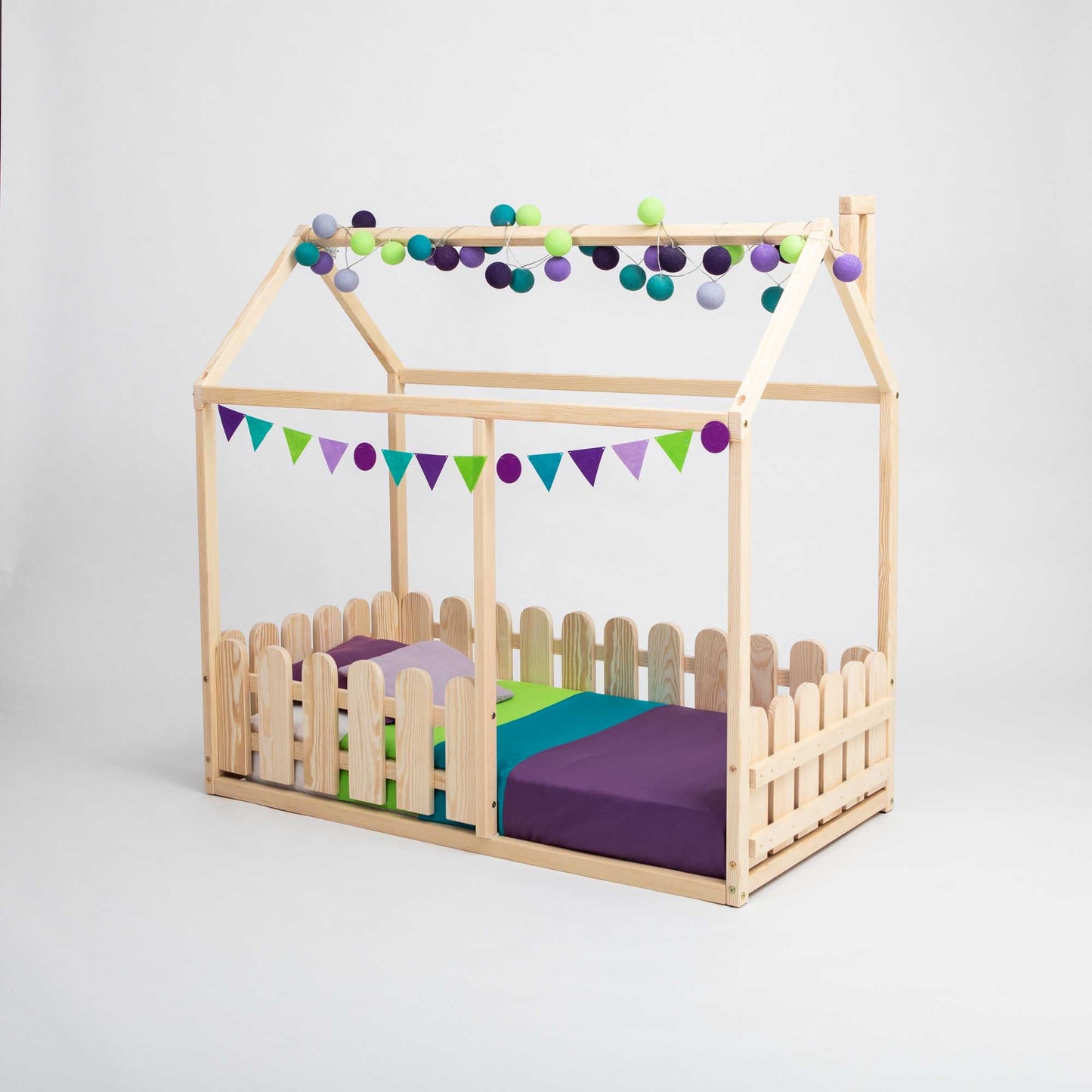 The Platform House Bed with a Picket Fence features a charming vertical picket fence design and comes complete with colorful bedding. It is adorned with a purple, green, and blue triangle banner along with a playful ball garland—making it an ideal choice for a toddler transitioning to their first big kid bed.
