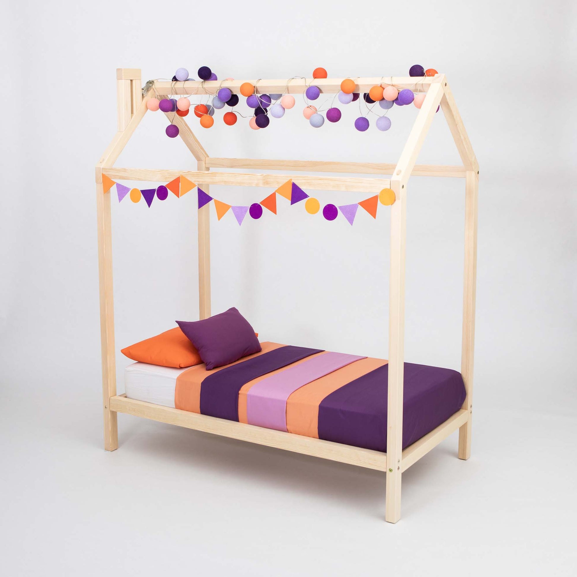 A cozy sleeping space featuring a *Wooden house bed on legs* decorated with colorful bunting and string lights in shades of orange, purple, and pink. The bed is made up with a purple and orange striped bedding set.