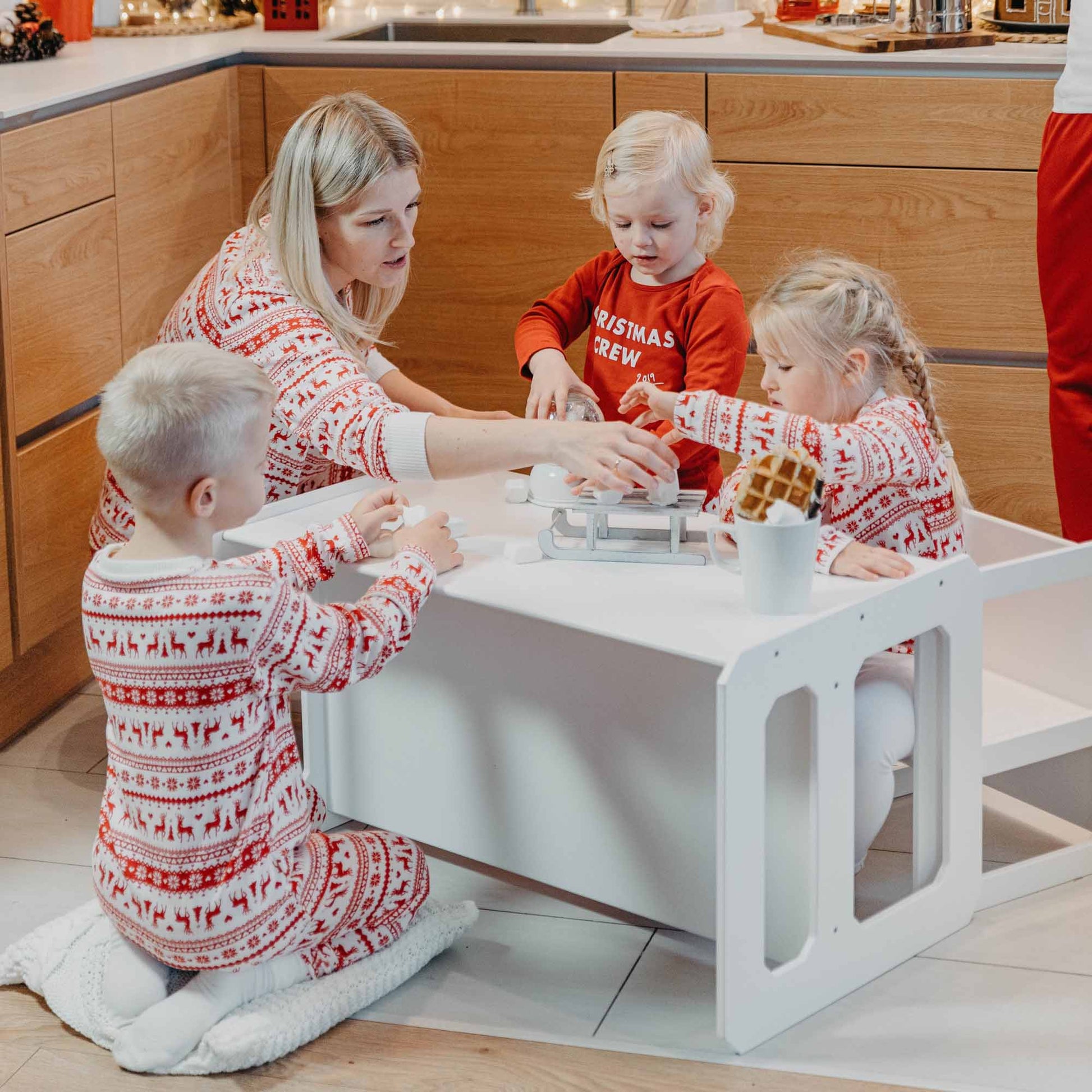 A woman and three children, all in Christmas-themed pajamas, are gathered around a Montessori weaning table and chair set in a kitchen setting, engaging in an activity together.