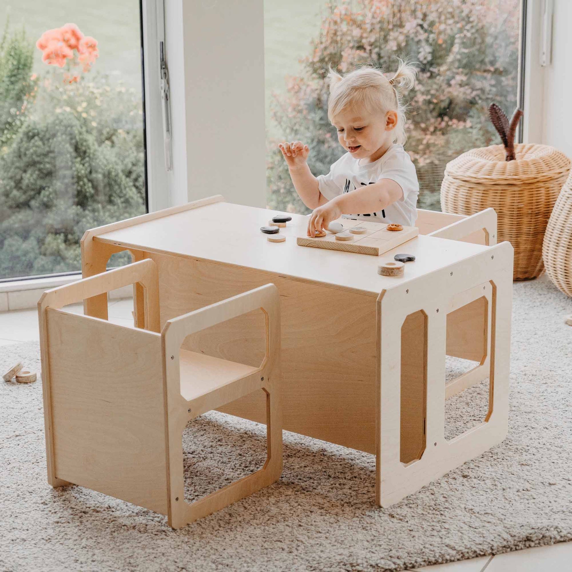 A young child engages with wooden toys at a Montessori weaning table and 2 chair set, fostering fine motor skill development in a room with large windows and a view of the garden.
