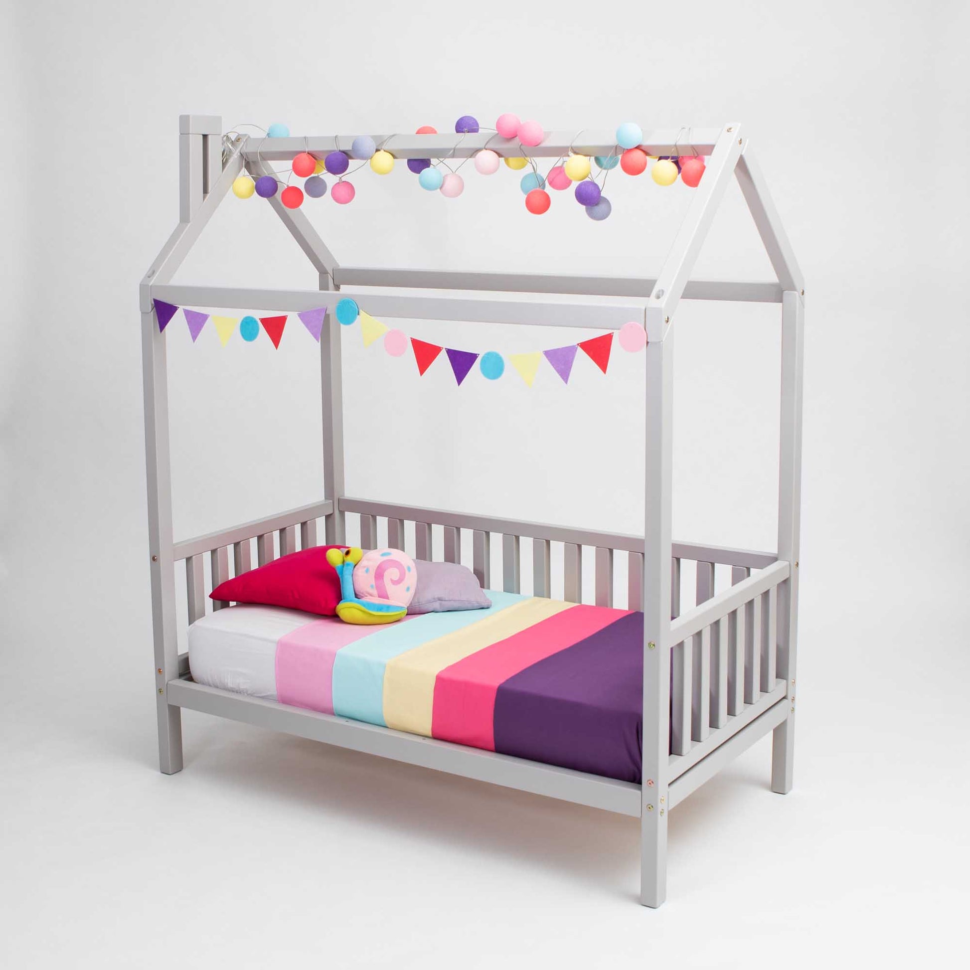 A raised house bed on legs with 3-sided rails, with a rainbow striped blanket and pom poms.