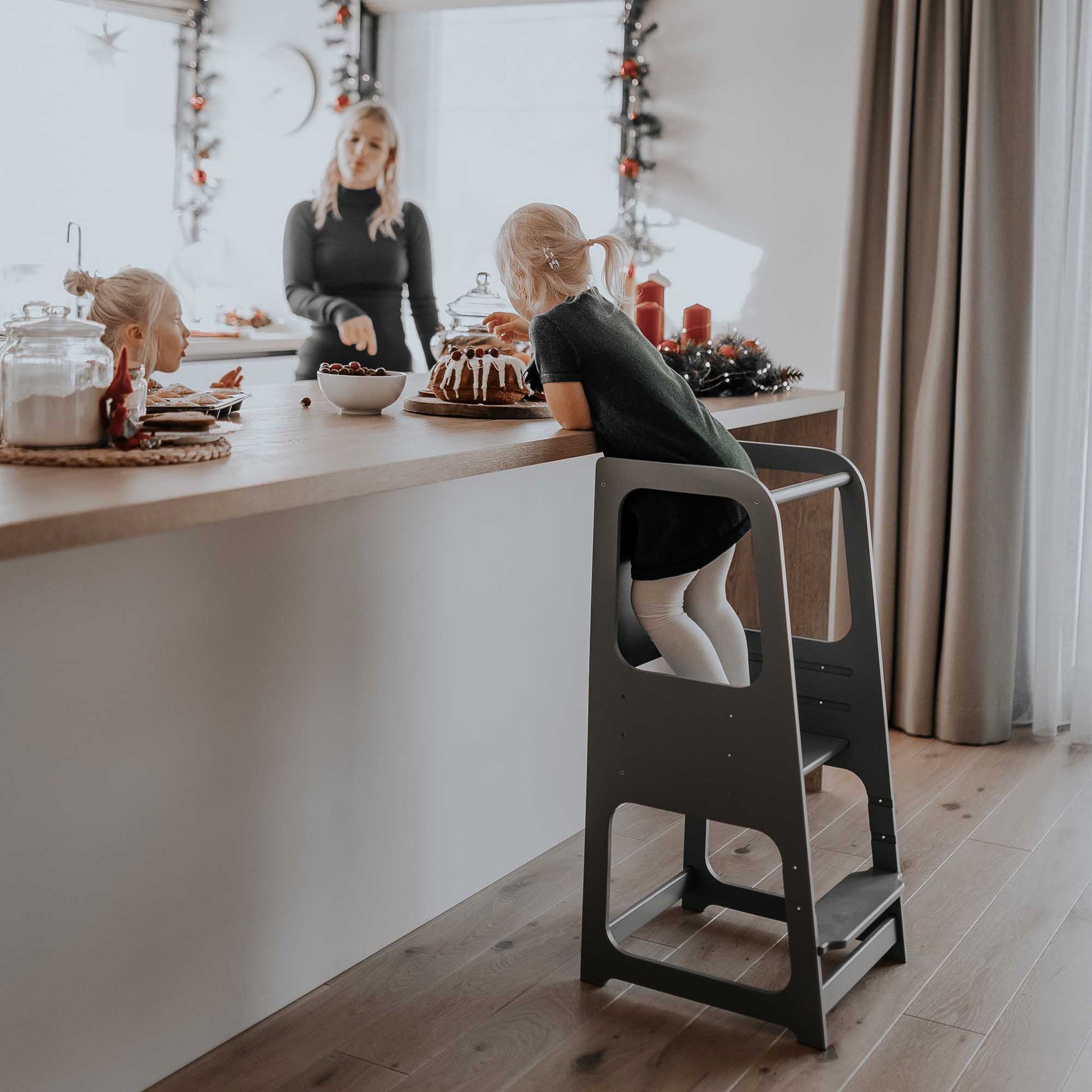 A toddler is standing on a Sweet Home From Wood Kitchen Tower with blackboard in a kitchen.