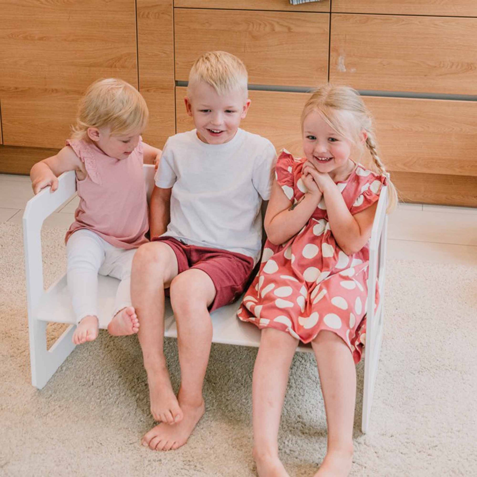Three children are seated on a white Montessori weaning table and chair set - two girls on either side of a boy. One girl is wearing a pink dress, the other has on a red polka-dot dress, and the boy is dressed in a white shirt and red shorts.