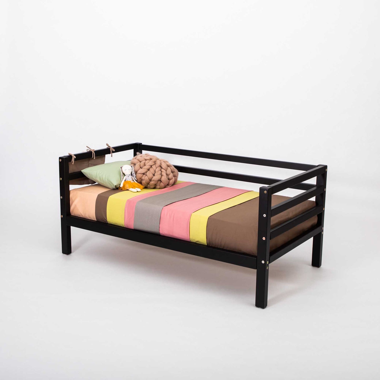 A Kids' bed on legs with a horizontal rail on 3 sides, crafted in small black solid wood, is styled with a colorful striped blanket, brown knot pillow, stuffed rabbit, and plush toy against a plain white background.