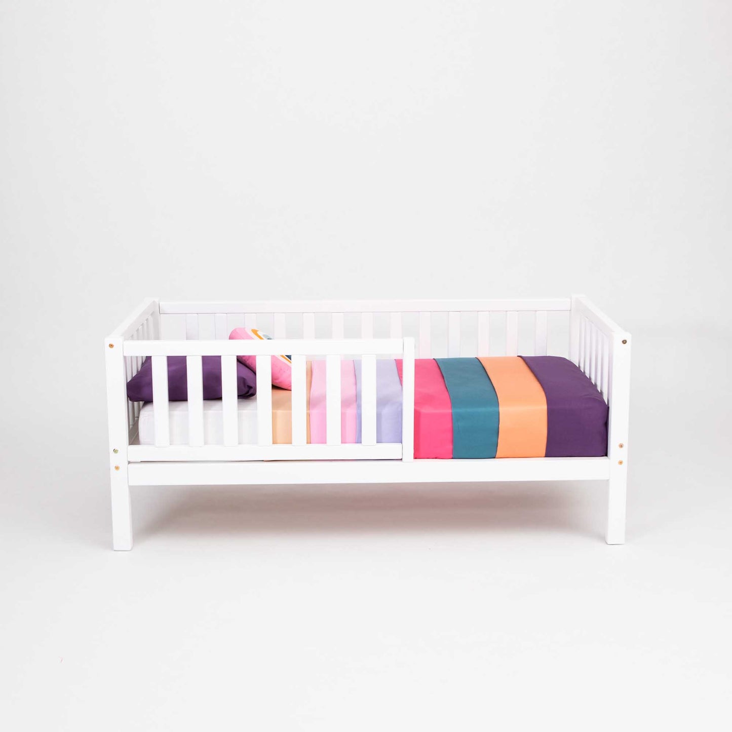 A Sweet Home From Wood toddler bed on legs with a fence in white, perfect for independent sleeping with a colorful striped sheet.