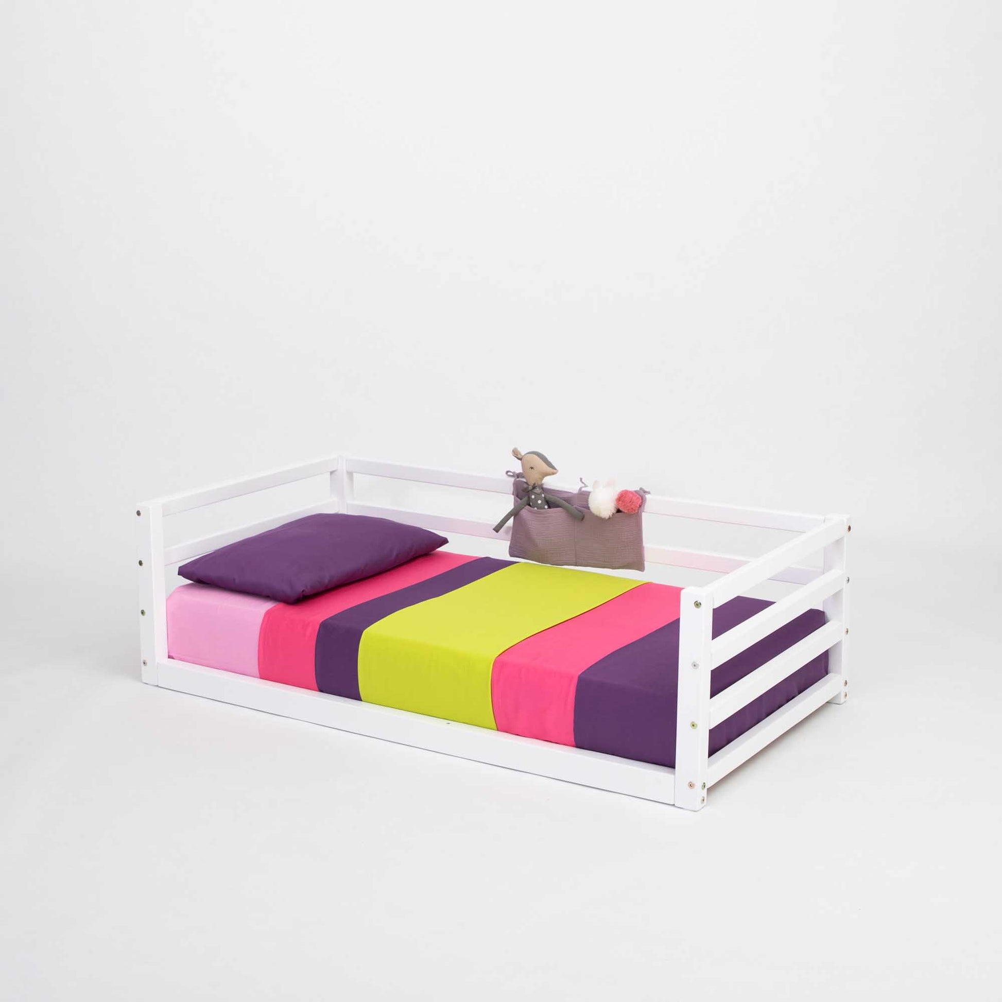 A Children's floor level bed with 3-sided safety rail by Sweet Home From Wood, with colorful sheets and a white frame, perfect for a toddler.