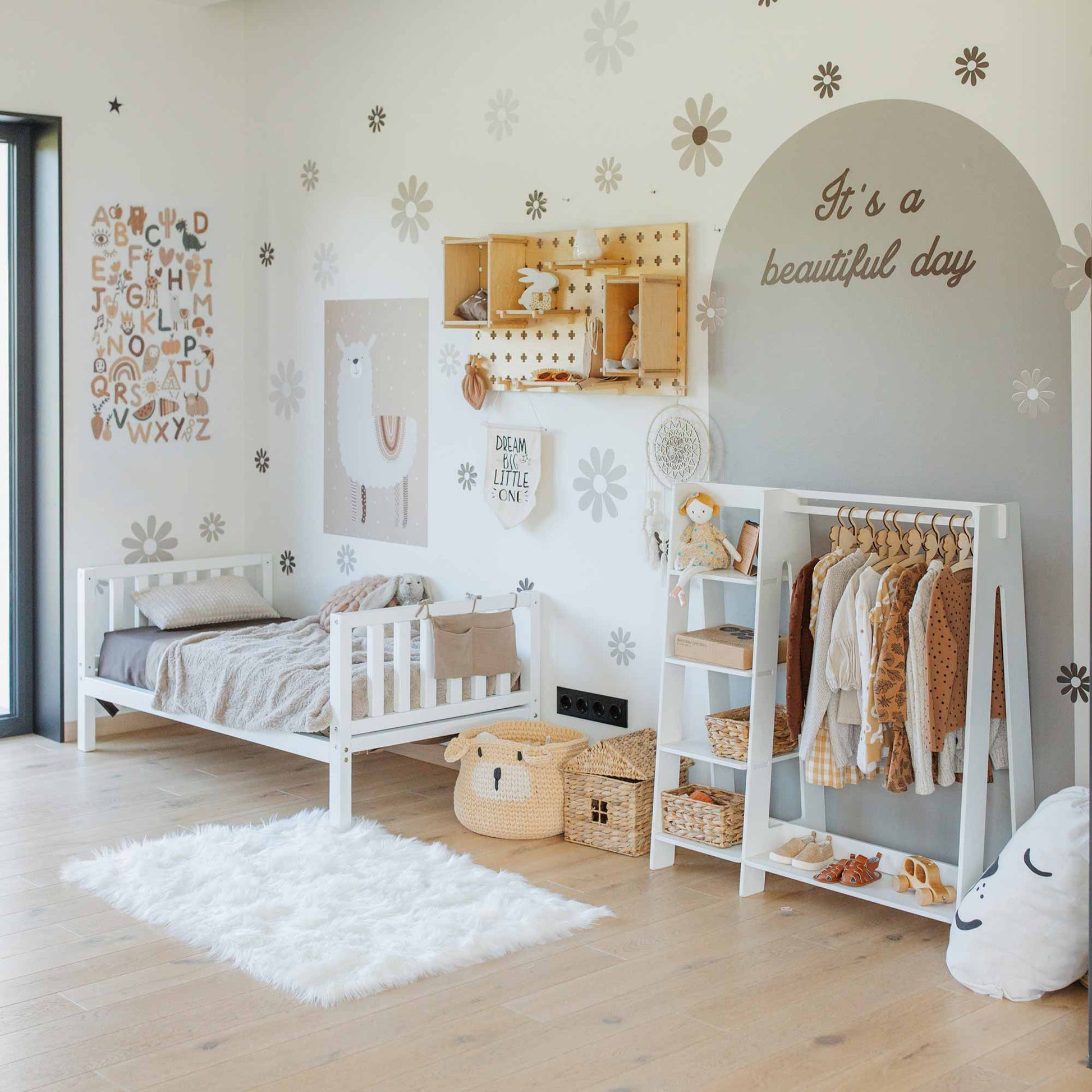 A neatly arranged children's room with a small bed, a Children's wardrobe with a dress-up clothing rack, Montessori-inspired storage shelves, and decorative items including wall art and baskets. The wall features the phrase "It's a beautiful day.