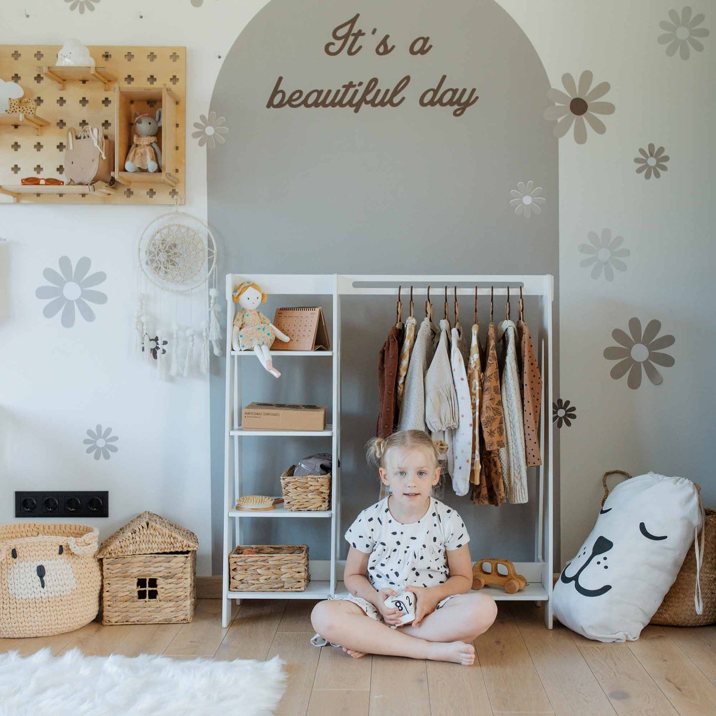 A child sits on a wooden floor in a Montessori-inspired room with a Children's wardrobe, dress up clothing rack, woven baskets, storage shelves lined with dolls, and a pillow. The wall behind features floral designs and the text "It's a beautiful day.