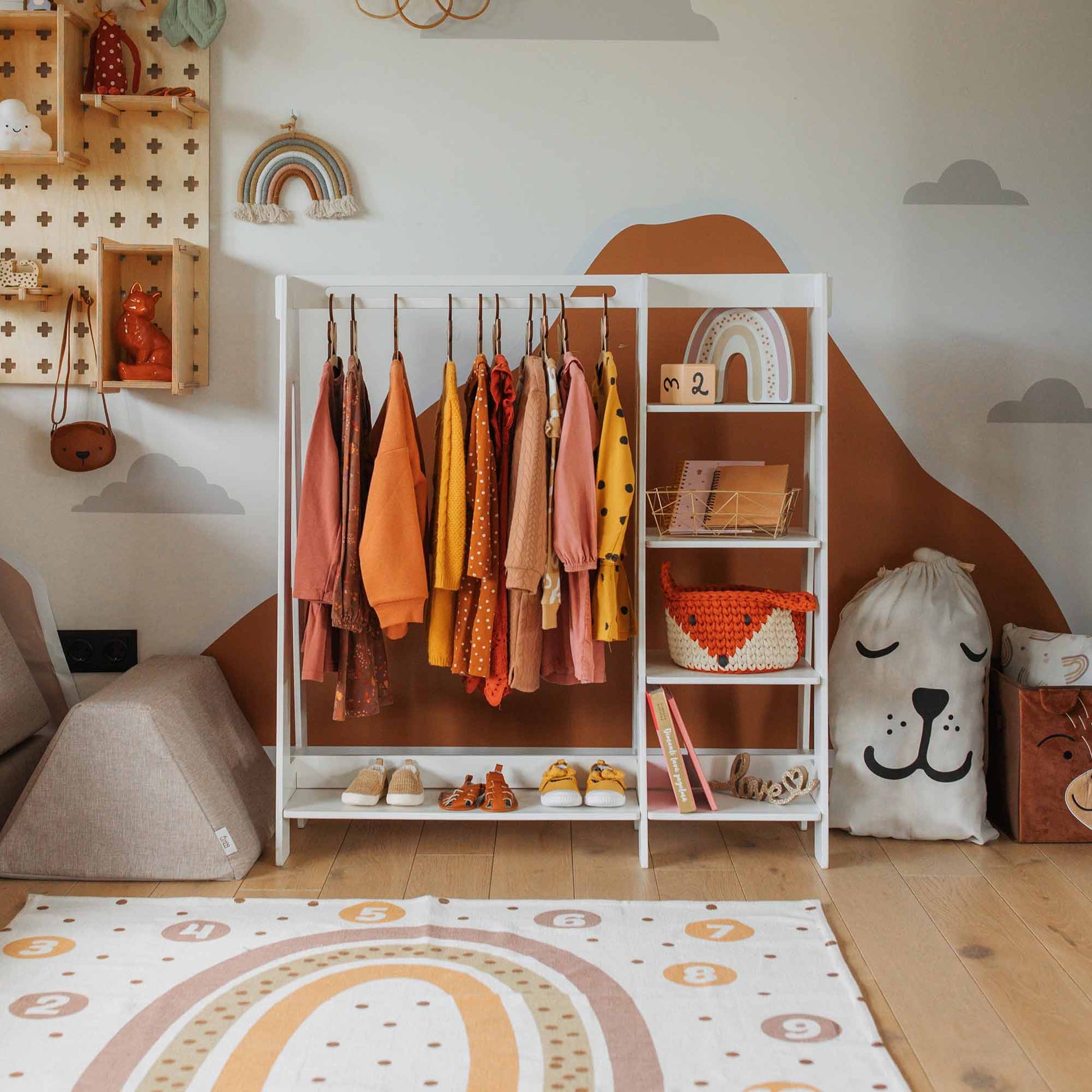 Children's room with a Montessori-inspired Children's wardrobe, dress-up clothing rack holding colorful clothes, storage shelves filled with books and toys, a large stuffed animal bag, and rainbow-themed decorations. A play mat featuring numbers on the floor adds an educational touch.