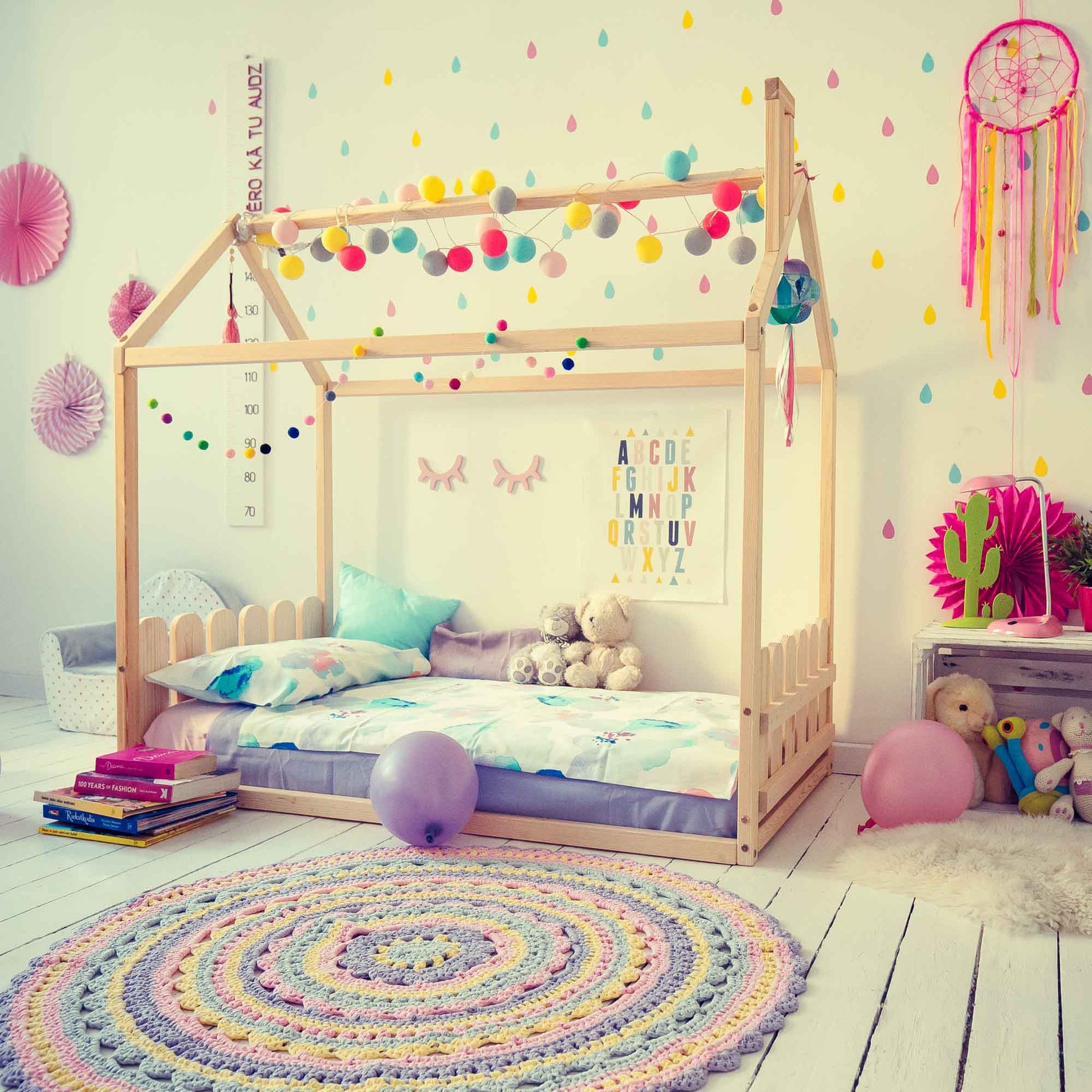 A child's bedroom featuring a Montessori house-frame bed with a picket fence headboard and footboard boasts colorful wall decorations, stuffed animals, pillows, and a crocheted rug on a white wooden floor.