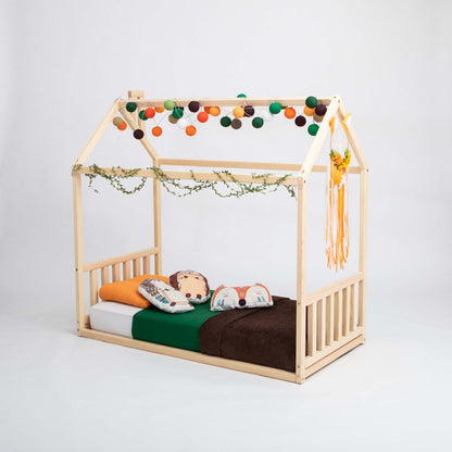 A Toddler house bed with a headboard and footboard, with a mattress, pillows, and blankets, decorated with string lights, green vines, and a hanging ornament on the side. Perfect as a preschool bed due to its zero-clearance design for added safety.