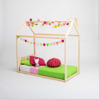 A Kids' house-frame bed with a picket fence headboard, designed to resemble a Montessori house bed, decorated with colorful string lights and bunting. The bed features a picket fence headboard, a green mattress, a pink pillow, and a red knitted cushion.