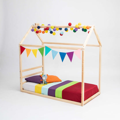 A Children's house bed with a horizontal headboard, decorated with colorful ball lights and rainbow bunting, features a red, green, yellow, and purple bedding set and a stuffed toy on the bed.