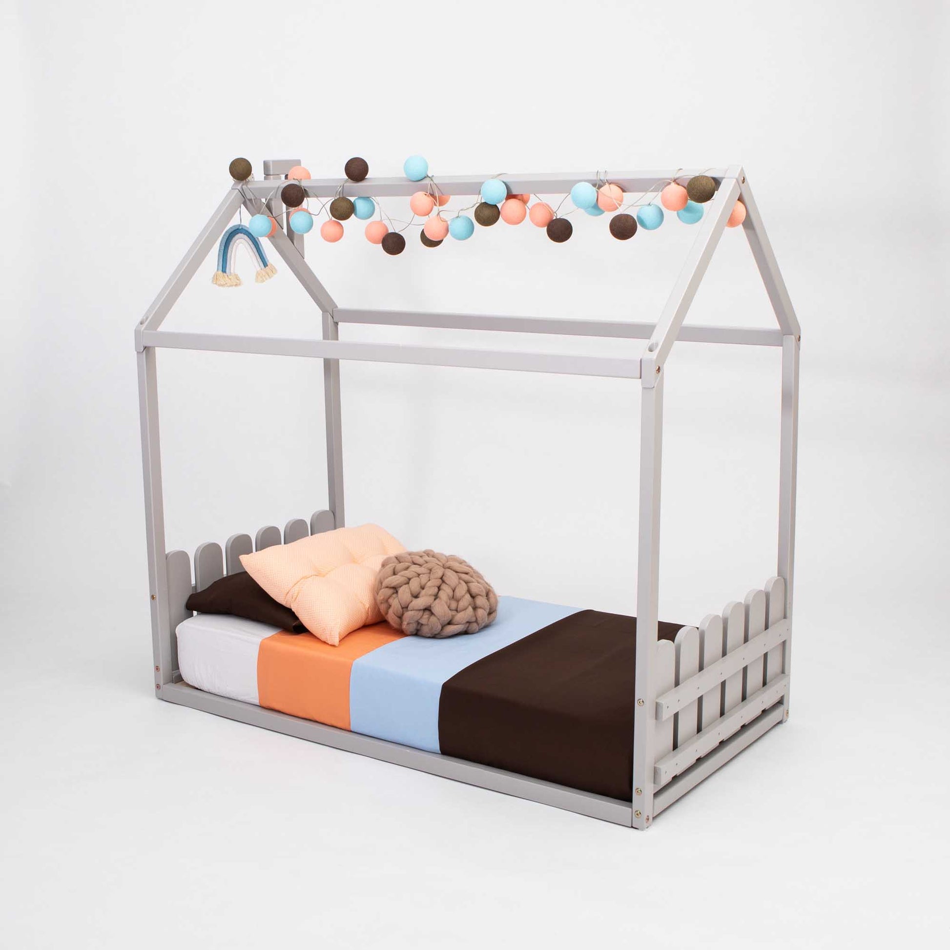 A Montessori house-frame bed with a picket fence headboard and footboard for toddlers with pom poms.