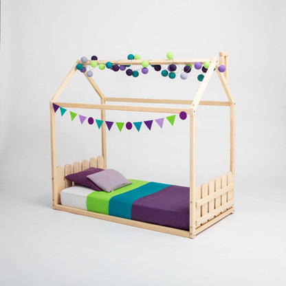 A Montessori house-frame bed with a picket fence headboard and footboard for kids with bunting and pom poms.