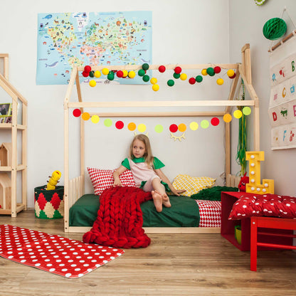 A little girl sits on a Montessori house-frame bed with a picket fence headboard and footboard in a playroom.