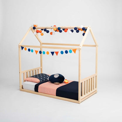 A Sweet Home From Wood toddler house bed with a headboard and footboard, featuring a wooden canopy and pom poms, creating a cozy sleep haven for children.
