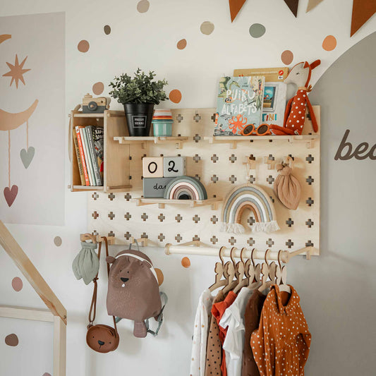 A children's room wall with a Pegboard with Clothes Hanger holding clothes, a backpack, and accessories. Adjustable shelves above display books, a plant, a clock, and rainbow decorations, enhancing the storage space while keeping everything organized and accessible.