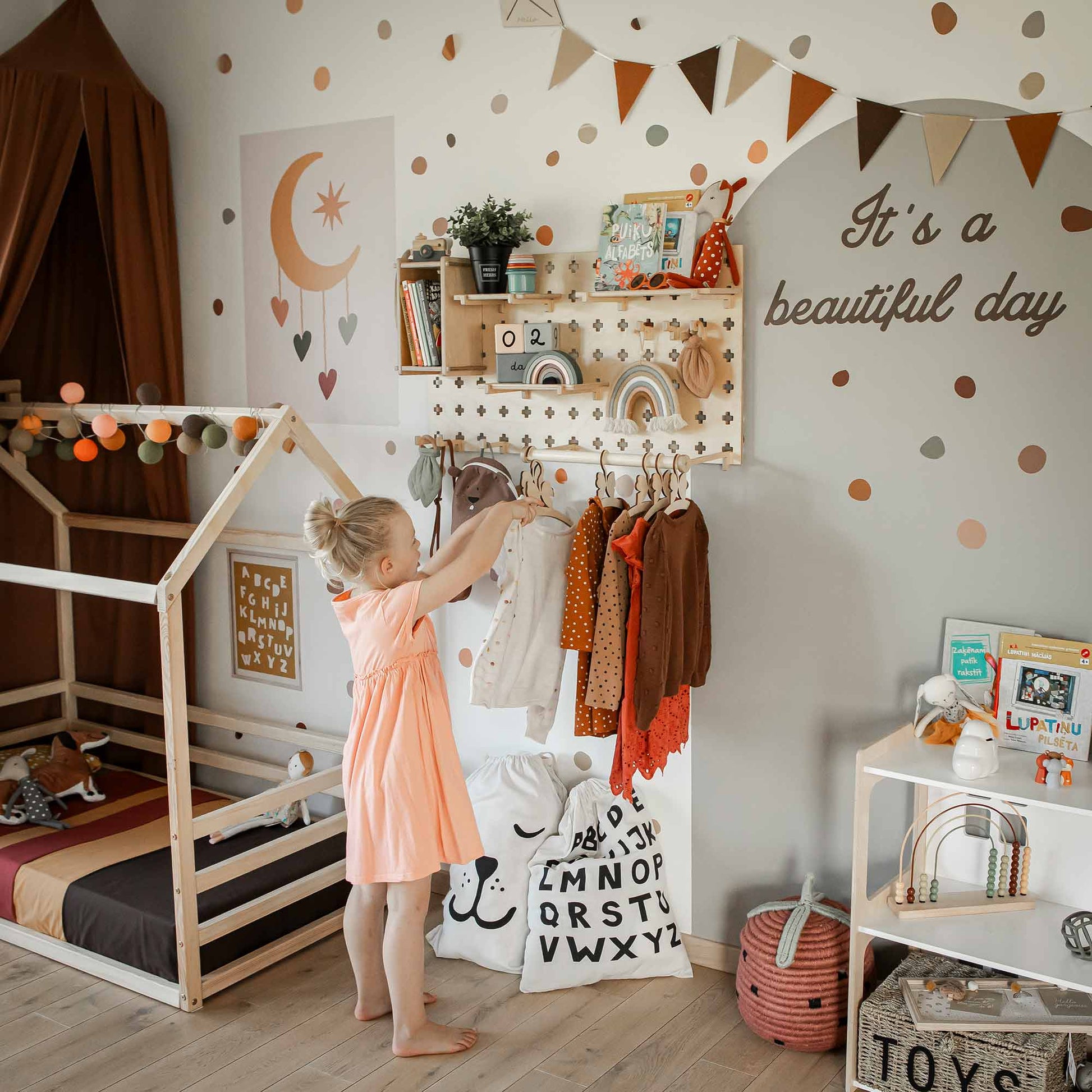 A young girl in a pink dress is reaching for clothes on a rack in a colorful, well-decorated children's room with wall decorations, toys, and a daybed. The room features a Pegboard with Clothes Hanger for extra storage space. The wall reads "It's a beautiful day.