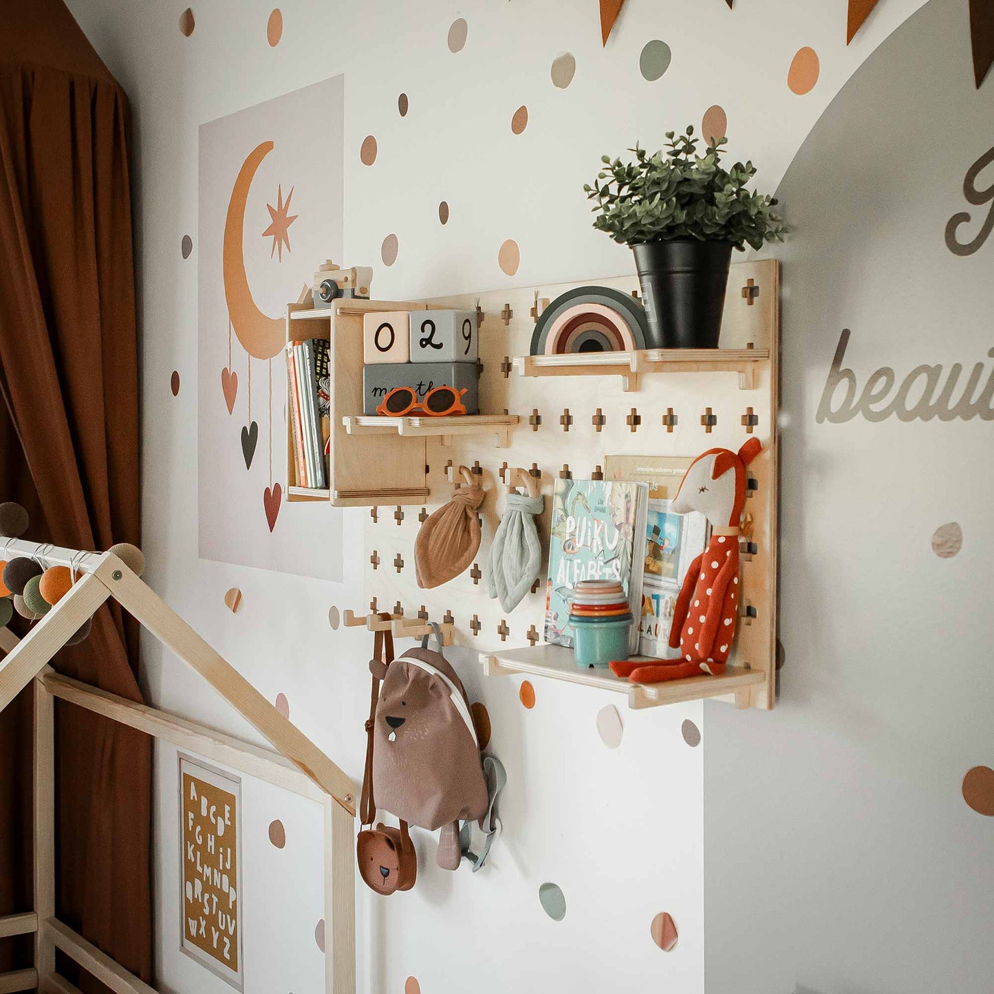 A child's room with a Floating Shelves Pegboard displaying toys, books, and a potted plant. The wall features a moon and stars art piece alongside a polka dot pattern. A customizable storage unit offers adjustable shelves beneath a partially visible wooden house-shaped frame.