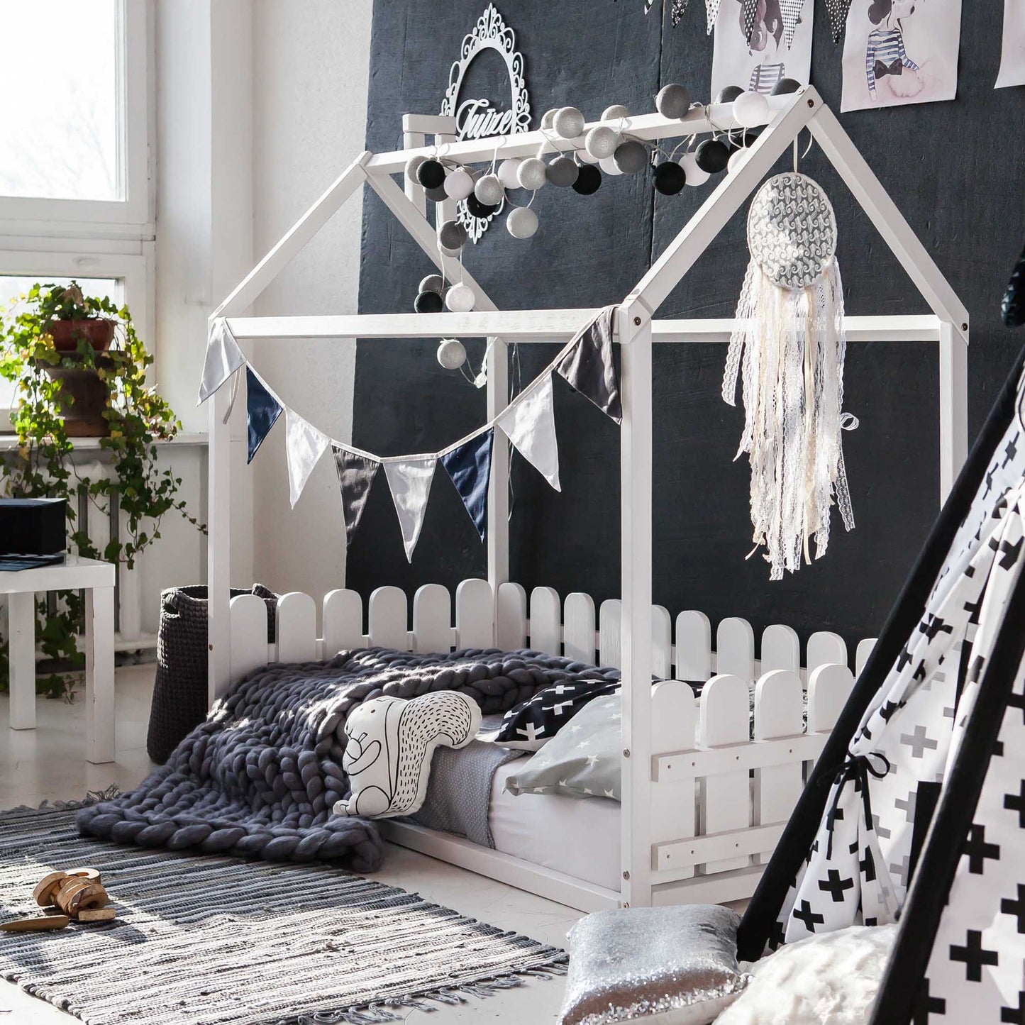 A child's room with a teepee, blankets, and pillows on a Floor house-frame bed with 3-sided picket fence rails.