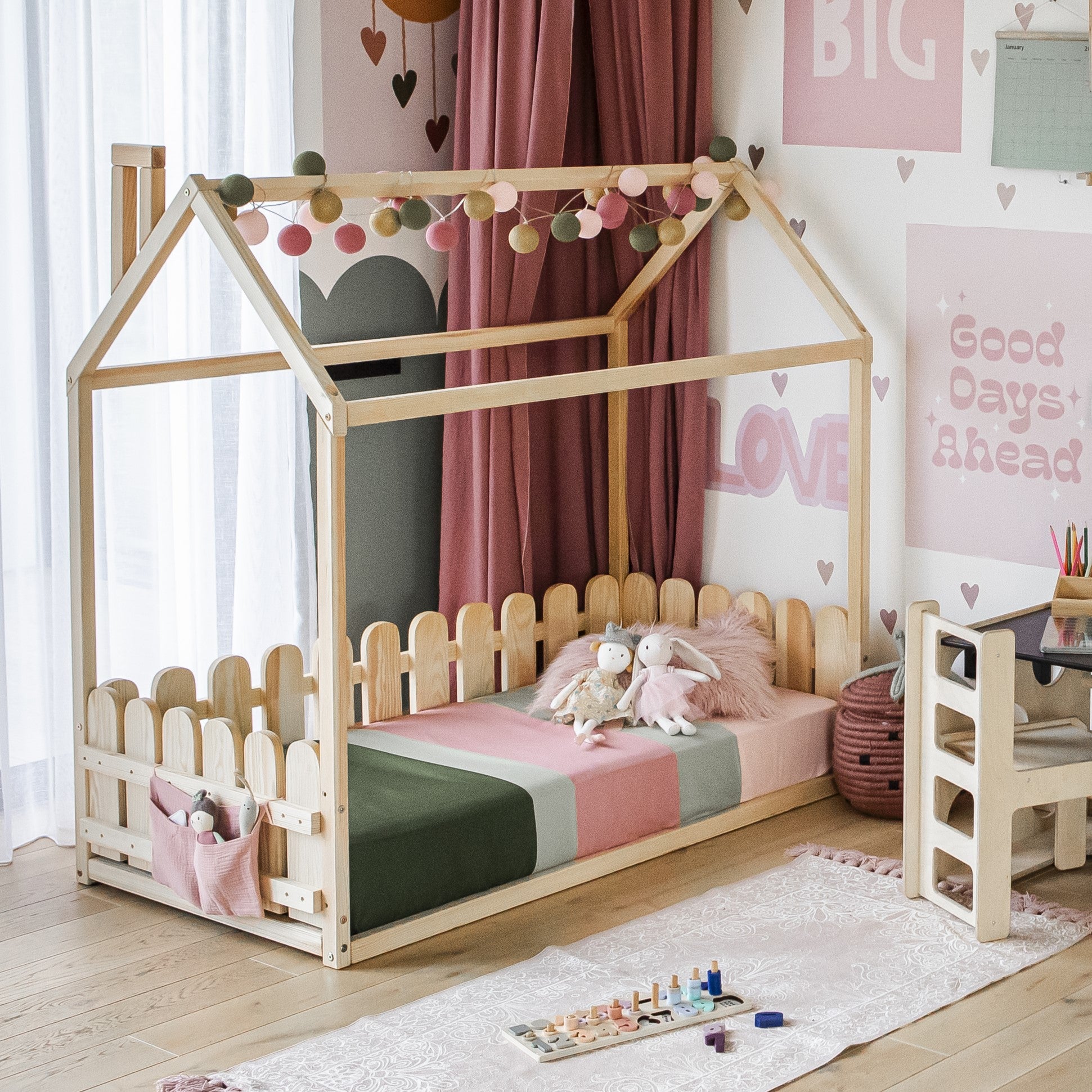 A child's bedroom featuring a Floor house-frame bed with 3-sided picket fence rails, adorned with pink and green bedding. The room is filled with stuffed animals and colorful decor. Motivational texts and hearts are displayed on the walls. Nearby, a small desk stocked with art supplies completes the cozy setting.