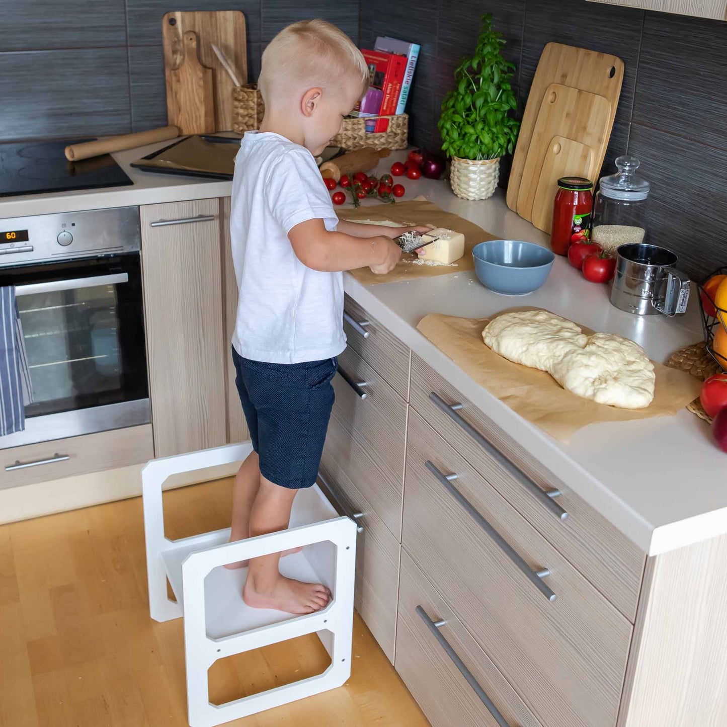 A young child sits at the Montessori weaning table and 2 chair set in a kitchen, independently cutting bread on the table next to dough and various kitchen items, showcasing fine motor skill development and fostering toddler independence.