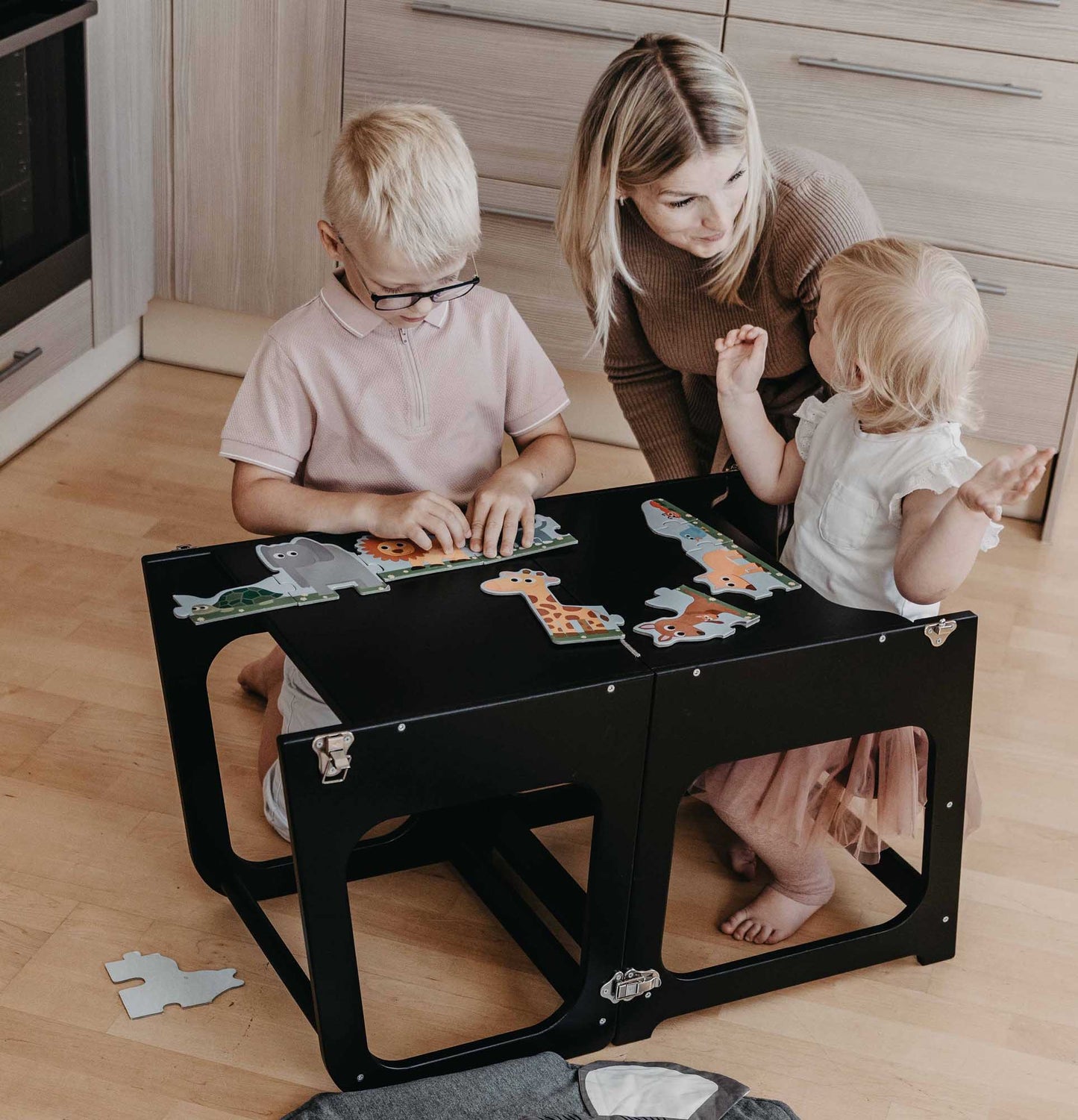 A woman and two kids playing with puzzles on a 2-in-1 Convertible kitchen tower - table and chair.