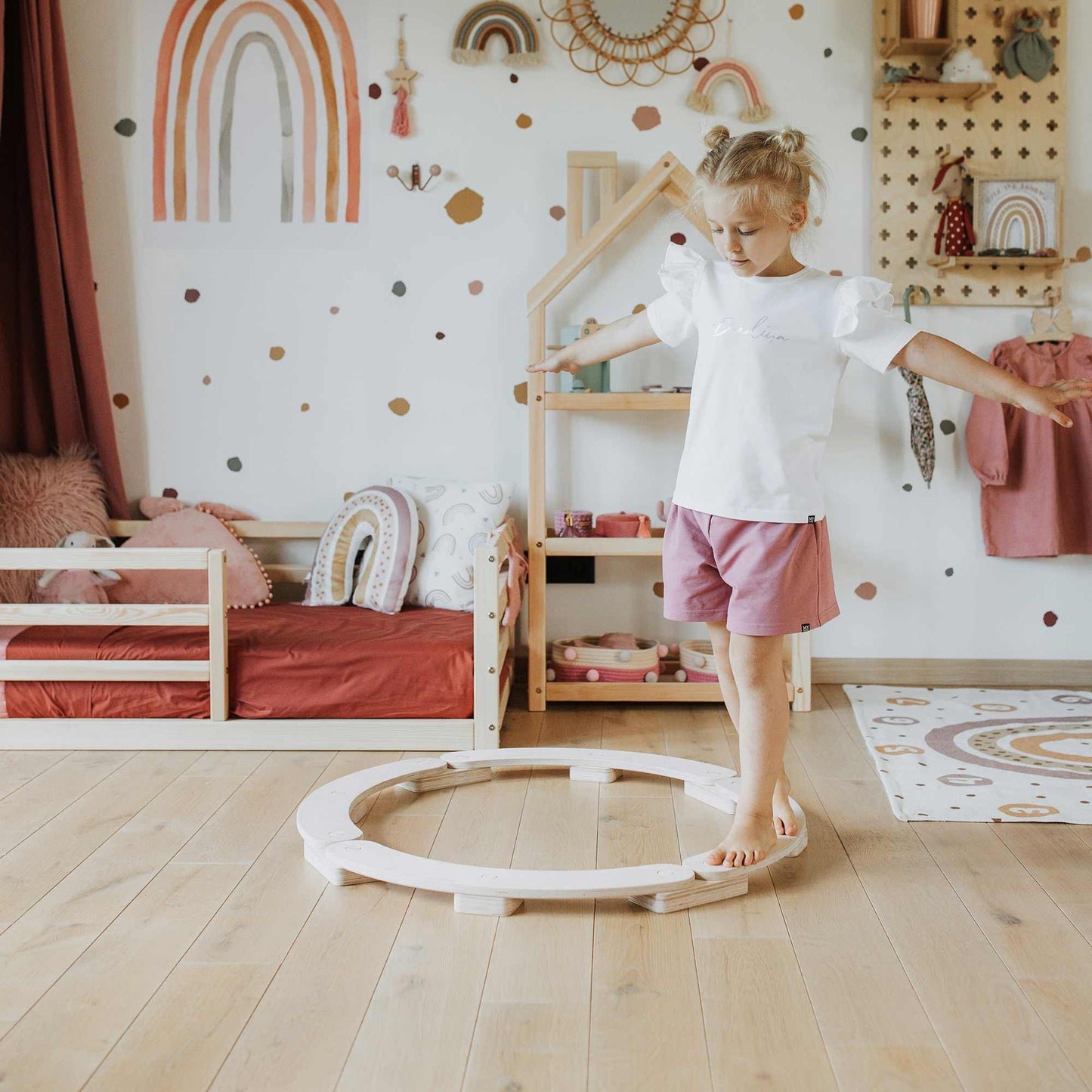 A child stands with arms outstretched on a circular Round Balance Beam, an essential component of the Balancing Beam Set, in a playroom decorated with pastel colors and rainbow motifs, enhancing their motor skills through this engaging Montessori indoor activity.