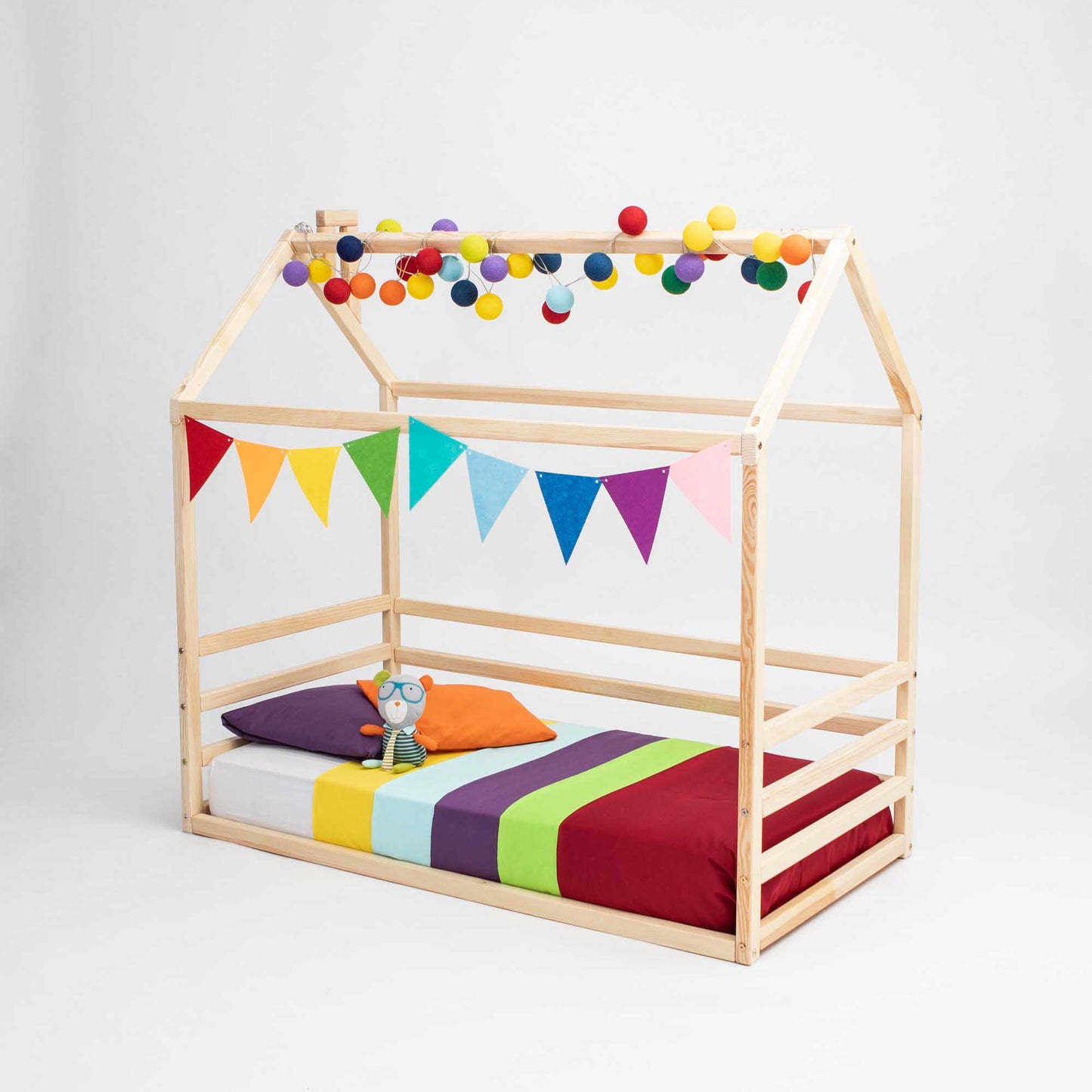 A Kids' house-frame bed with 3-sided horizontal rails features a colorful striped mattress, pillows, a plush toy, triangular pennant banners, and multi-colored pom-pom garlands.