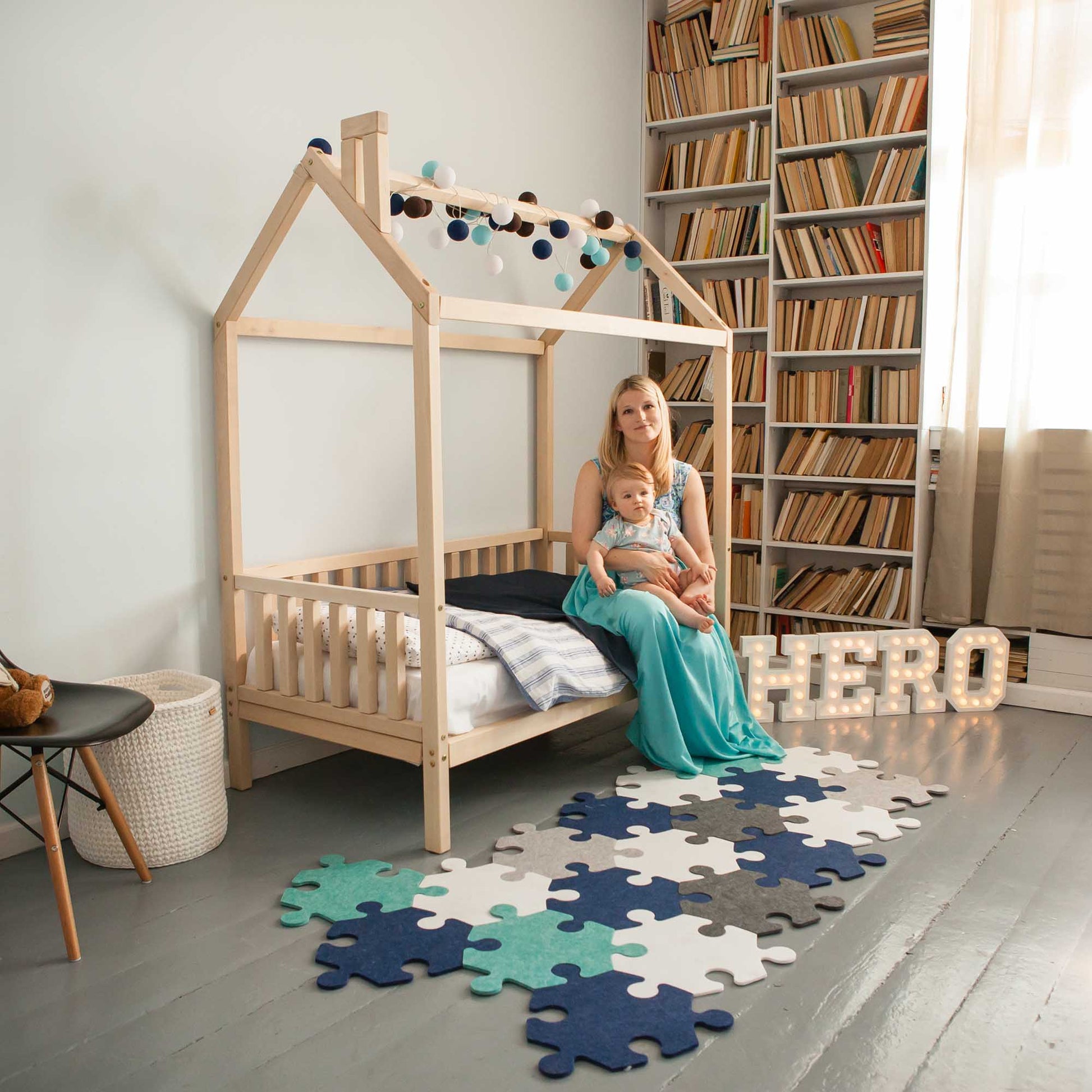 A woman with a baby in a room with a raised house bed on legs with 3-sided rails and bookshelves.