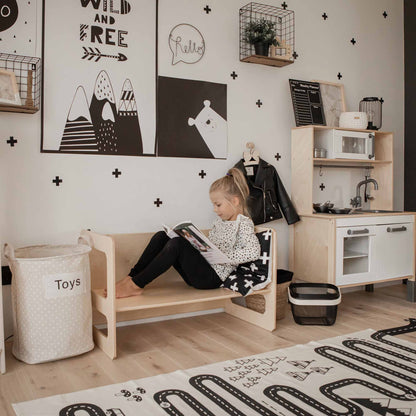 A child sits on a wooden bench reading a book in a monochromatic playroom with toy storage, a play kitchen, and wall art featuring mountains and animals. Nearby, the Montessori weaning table and 2 chair set fosters fine motor skill development and encourages toddler independence.