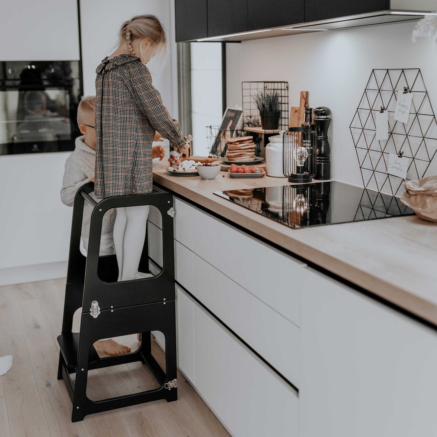 A child is standing on a Sweet Home From Wood 2-in-1 transformable kitchen tower - table and chair in a kitchen.
