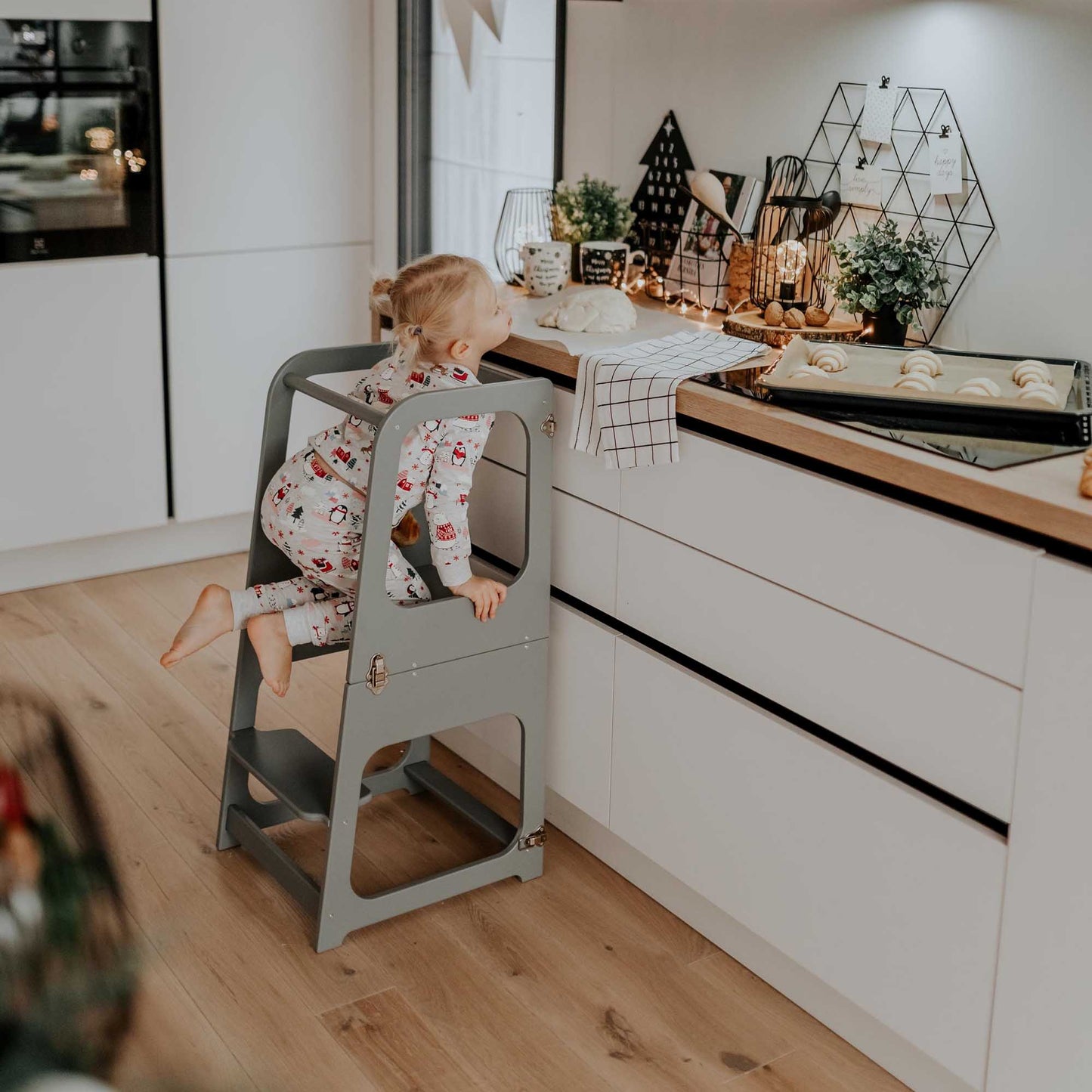 A toddler in pajamas standing on a 2-in-1 Convertible kitchen tower - table and chair in a kitchen.