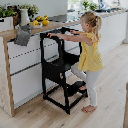 A toddler standing on a 2-in-1 Convertible kitchen tower - table and chair in a kitchen.