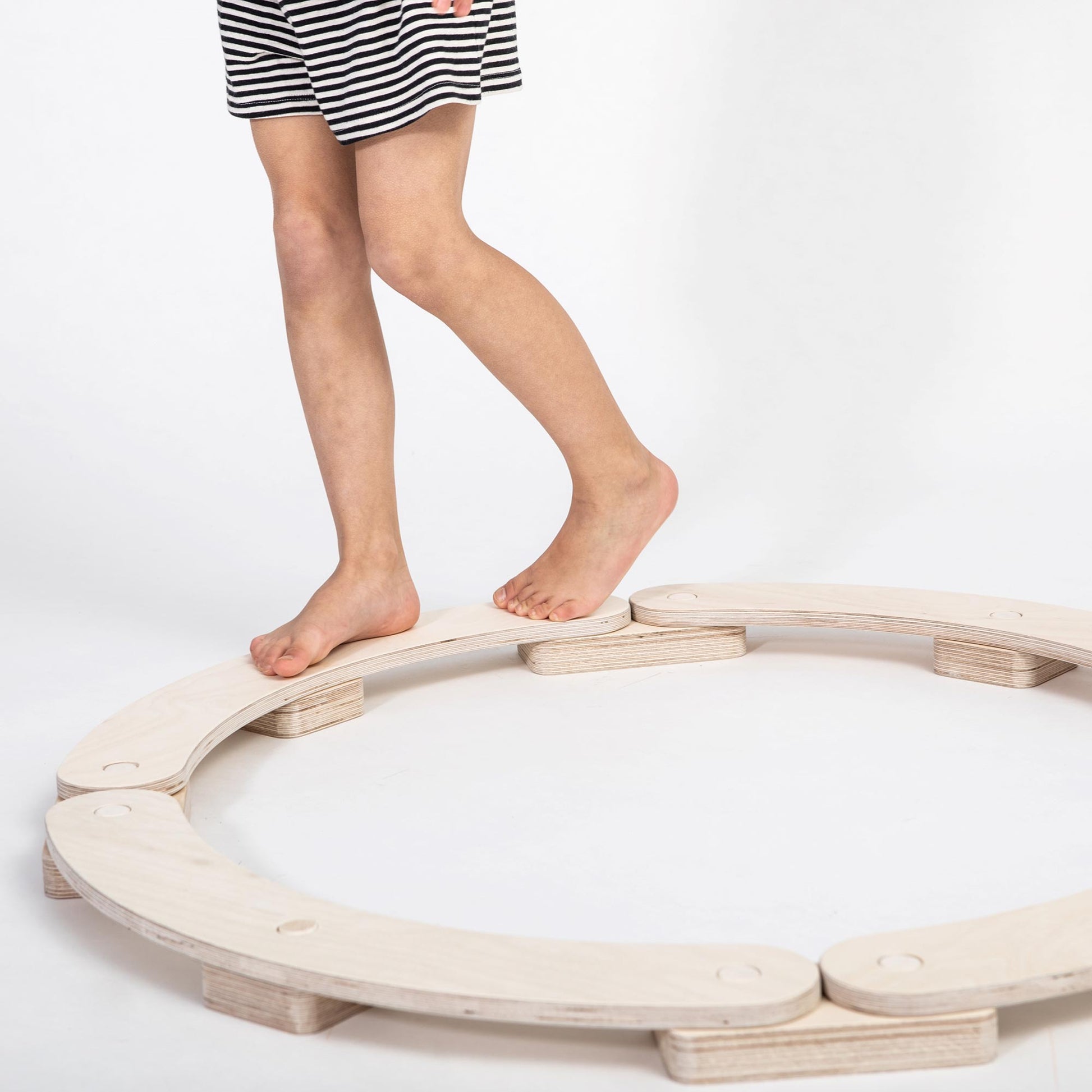 A child in striped shorts balances barefoot on a circular Round Balance Beams set, an excellent Montessori indoor activity for developing motor skills, set against a white background.