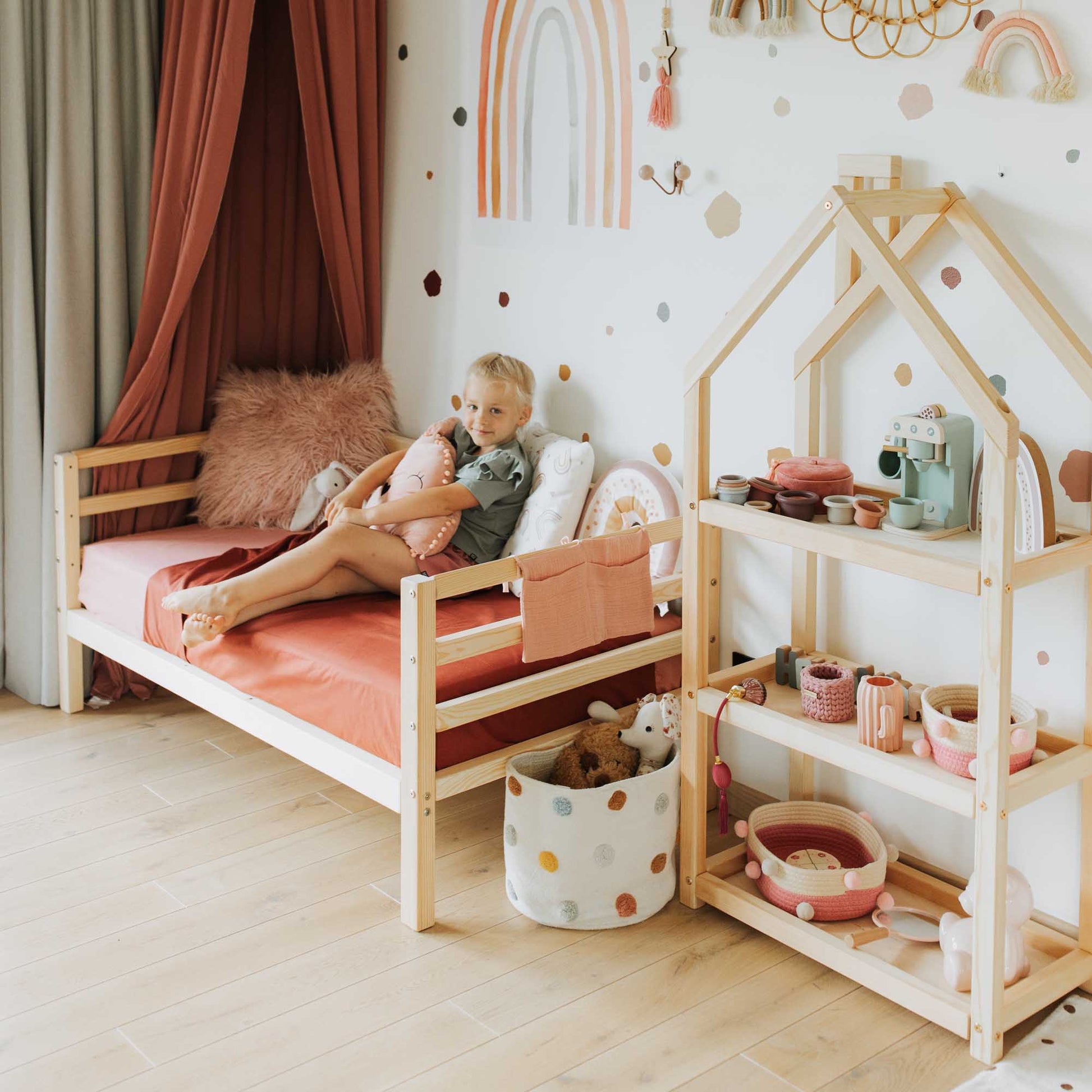 A girl is sitting on a Sweet Home From Wood 2-in-1 transformable kids' bed with a 3-sided horizontal rail in a child's room.