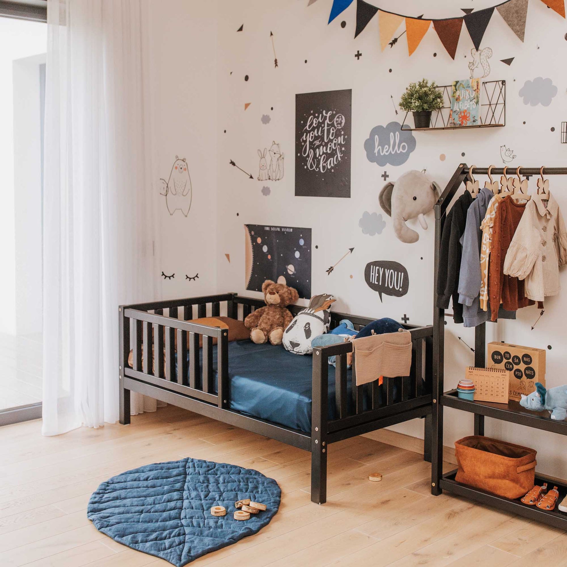 A children's room with a Sweet Home From Wood 2-in-1 toddler bed on legs with a vertical rail fence, shelves, and a teddy bear.