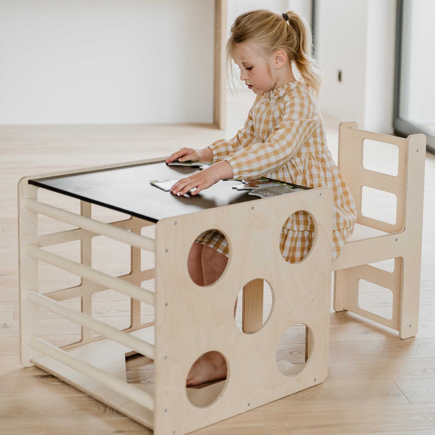A little girl sitting at a Sweet Home From Wood wooden desk with a Sweet Home From Wood tablet, Climbing arch + Transformable climbing cube / table and chair  + a ramp.