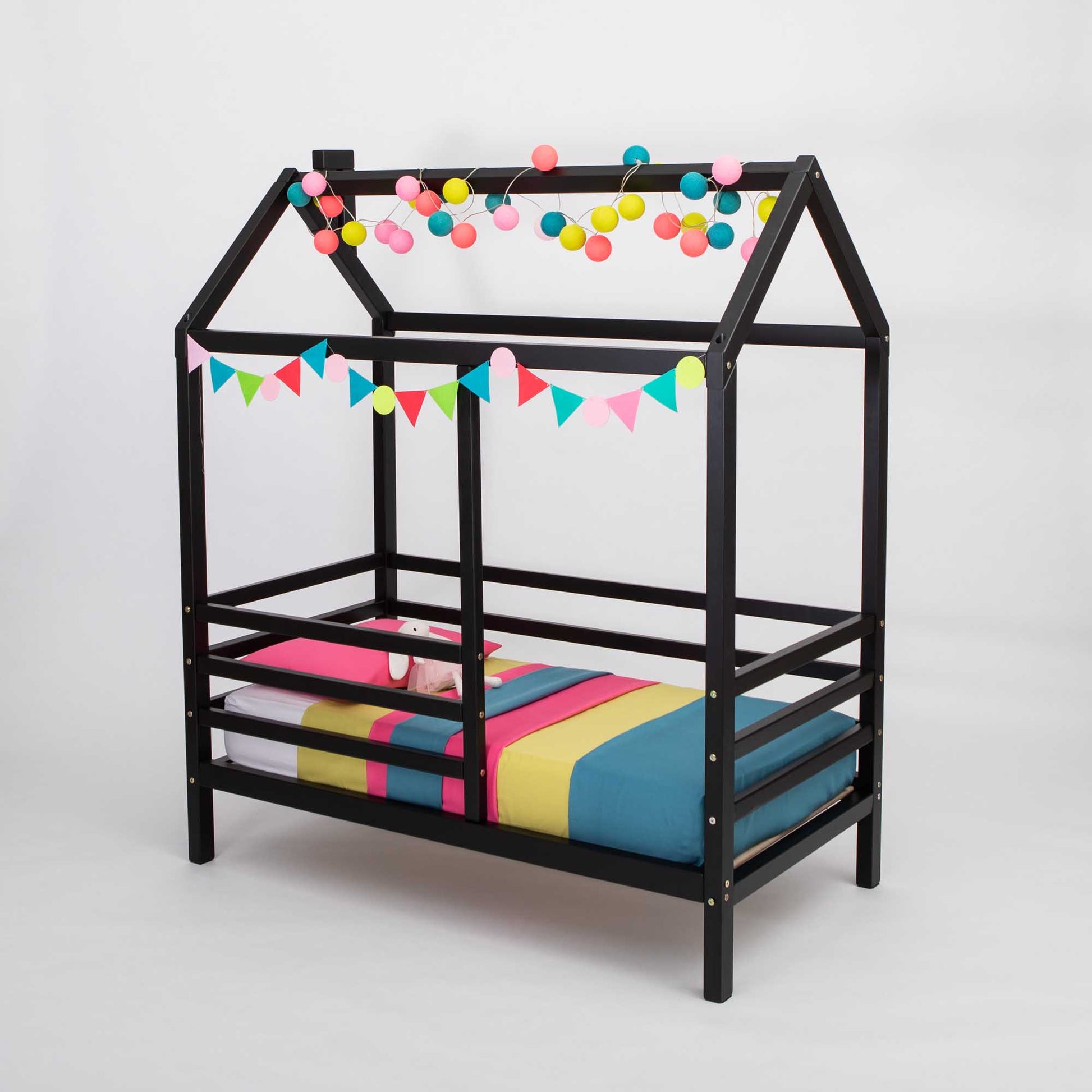 A wooden house bed on legs with a fence decorated with colorful pom poms.